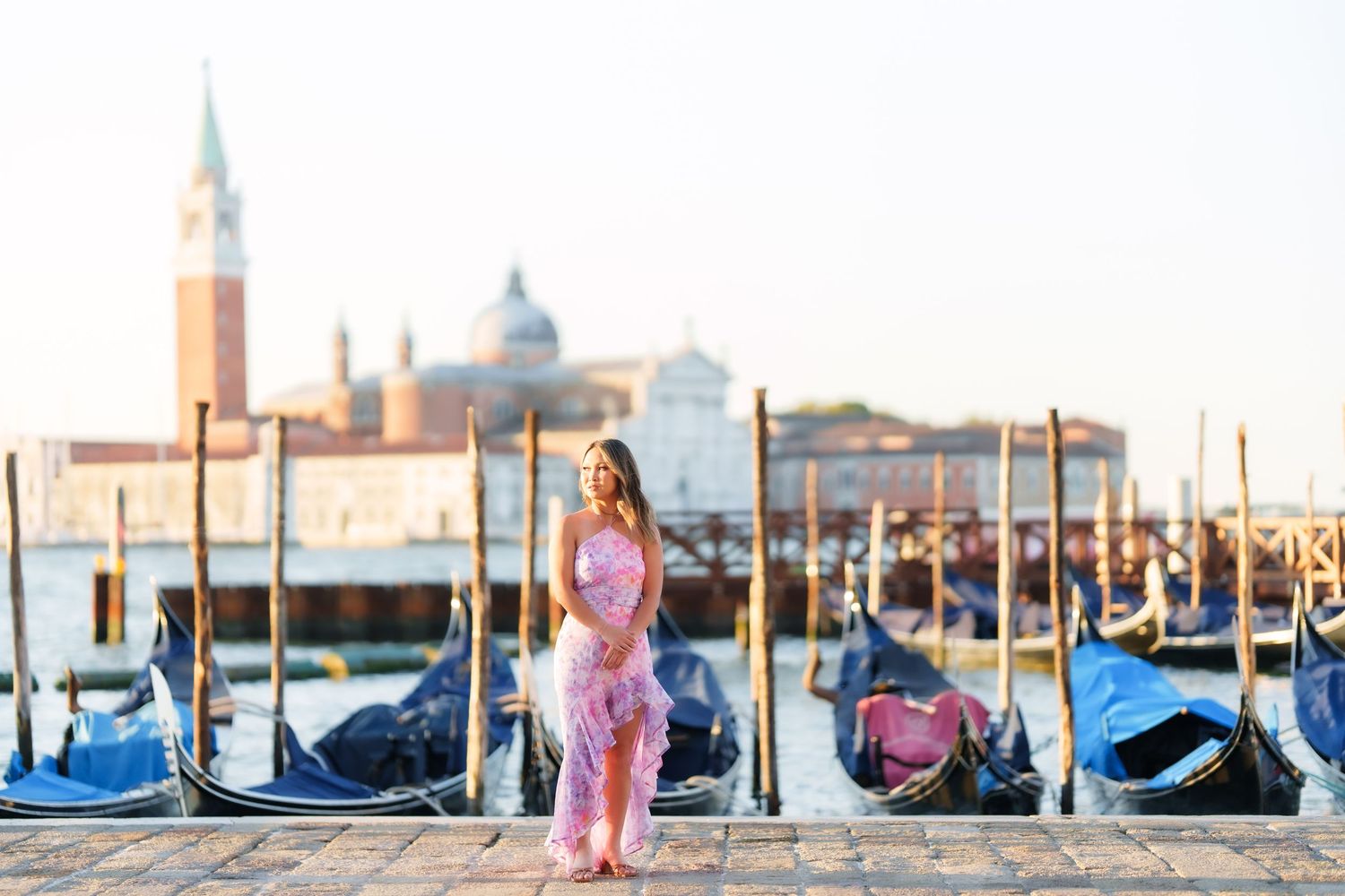 Woman in pink dress standing by Venice gondolas with San Giorgio Maggiore church in background at sunrise.