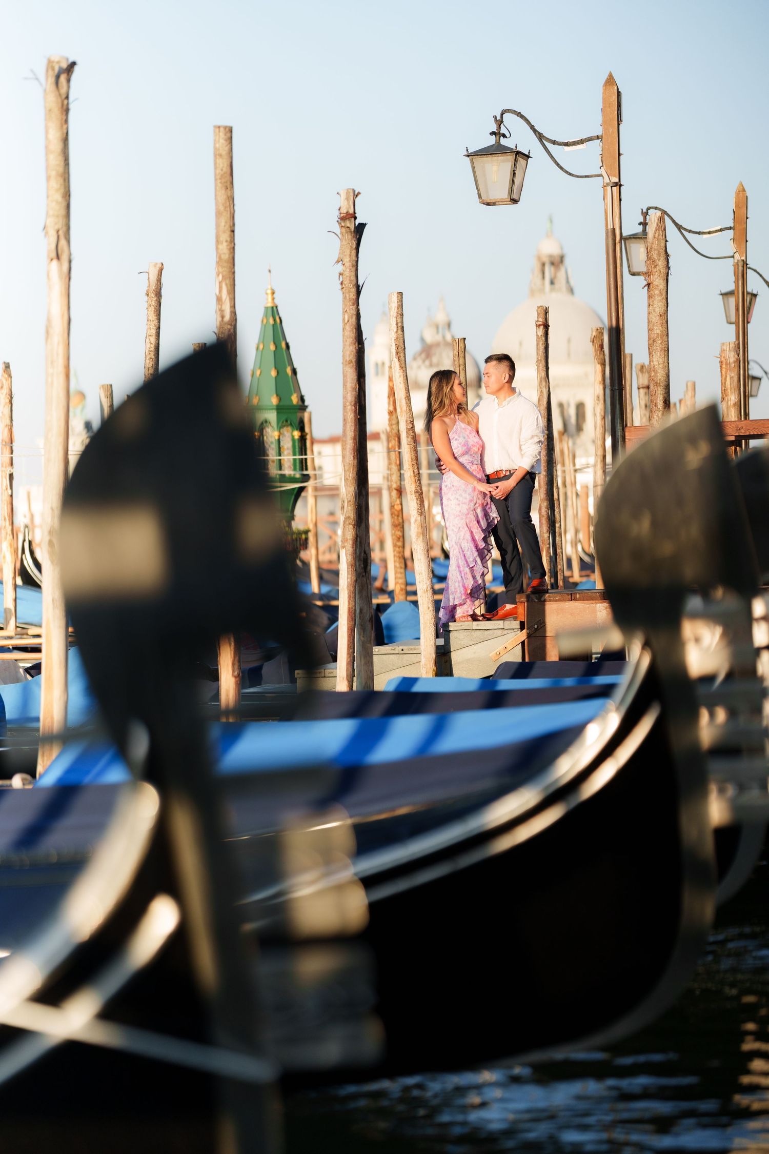 Couple embracing among gondolas and wooden posts at Venice waterfront with San Giorgio Maggiore in background.