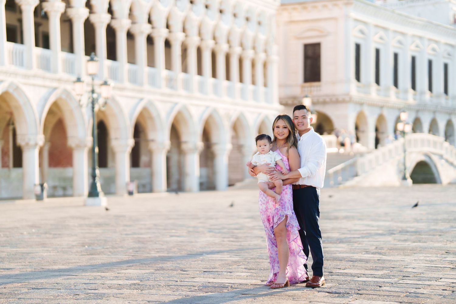 Family posing in St. Mark's Square Venice, mother in pink dress holding baby, father beside her, Doge's Palace backdrop.