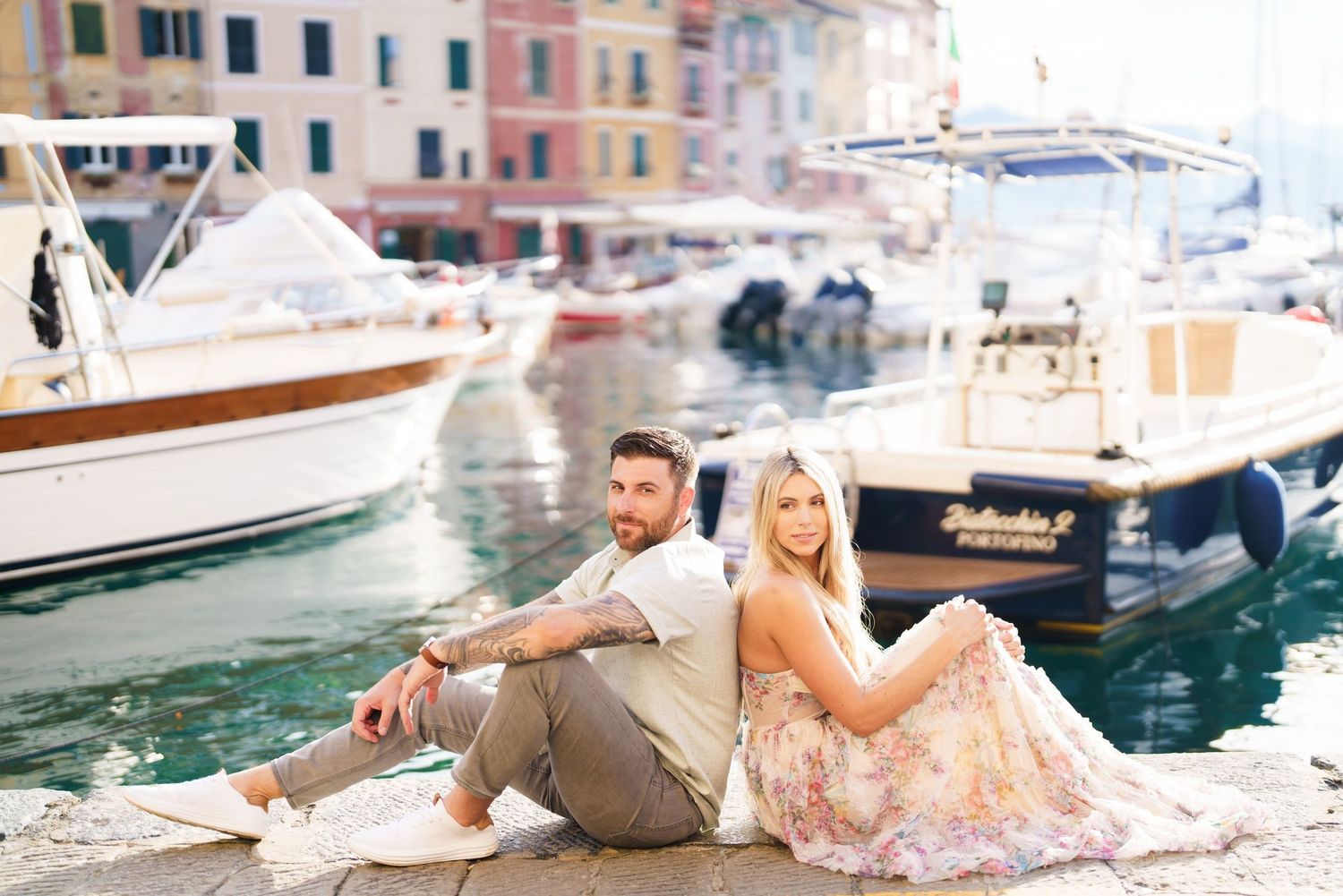 Couple sitting back-to-back on a dock in Portofino, Italy, with colorful boats and waterfront buildings behind them.