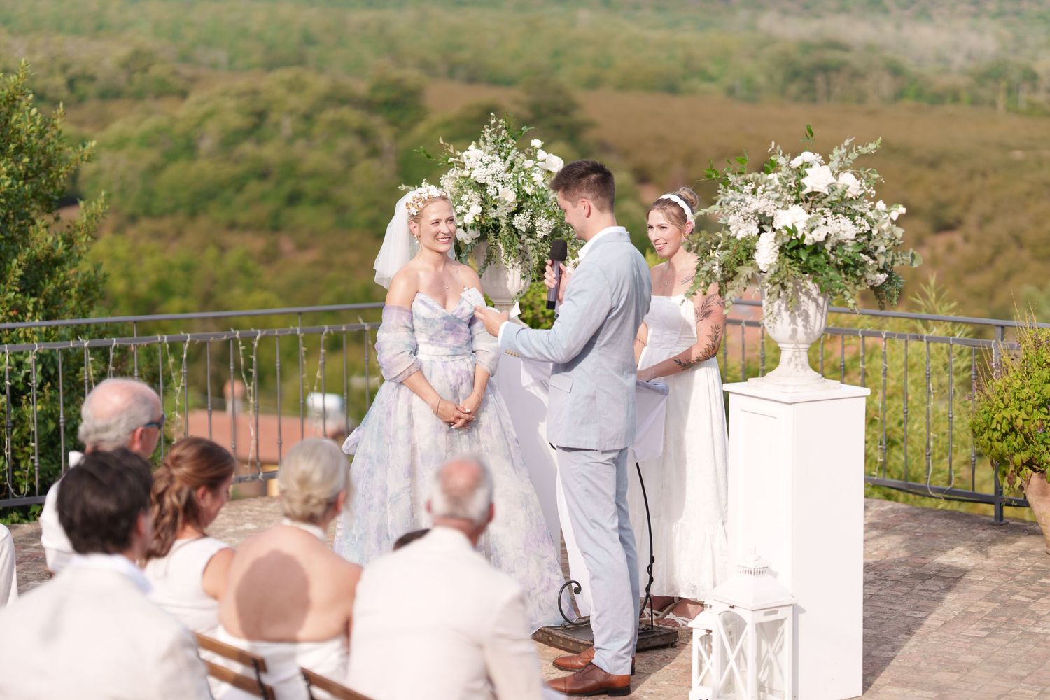 Outdoor wedding ceremony on a terrace with bride and groom exchanging vows, white floral arrangements, and scenic greenery backdrop.