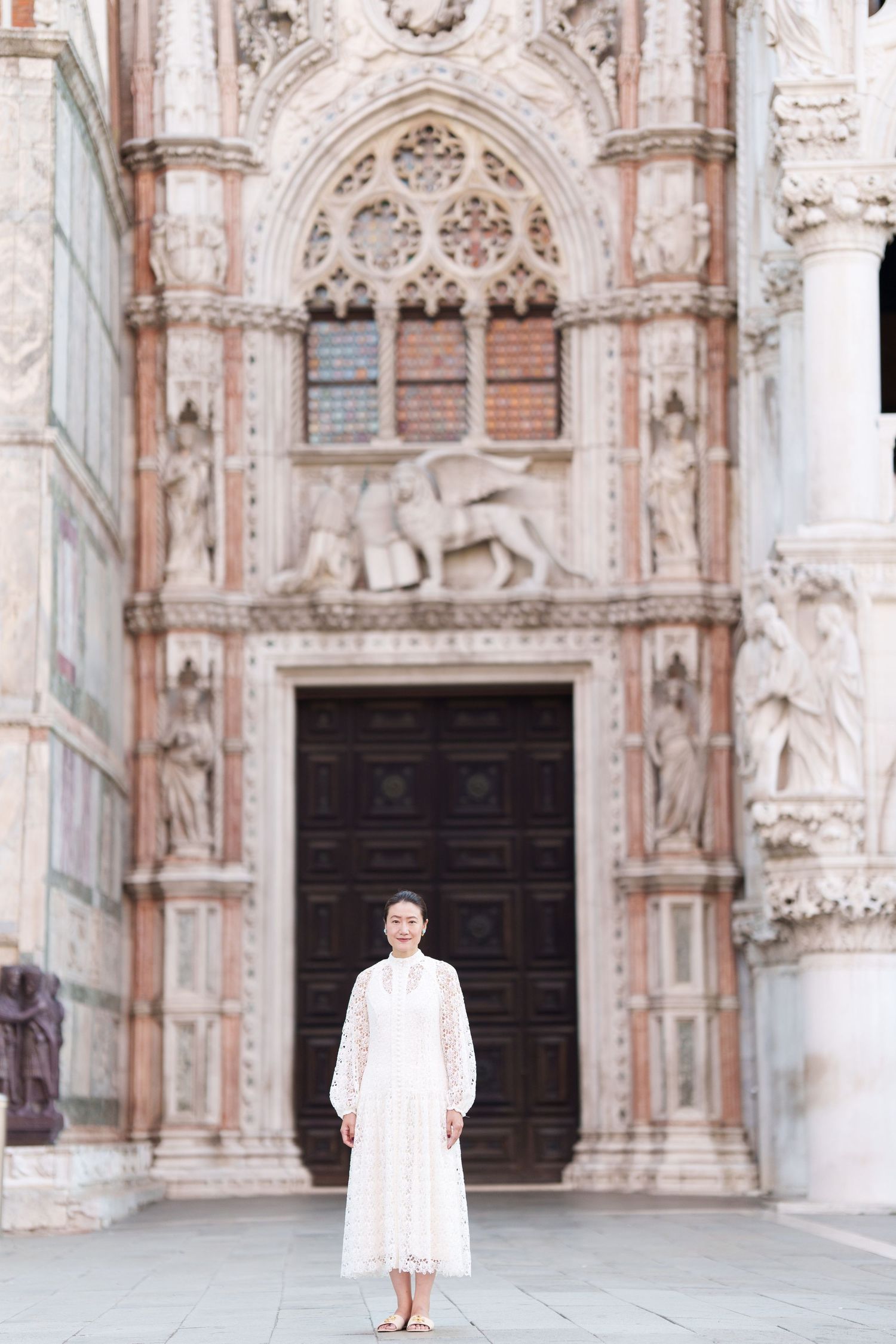 Woman in white lace dress standing before ornate Gothic cathedral entrance with marble sculptures and large wooden doors.