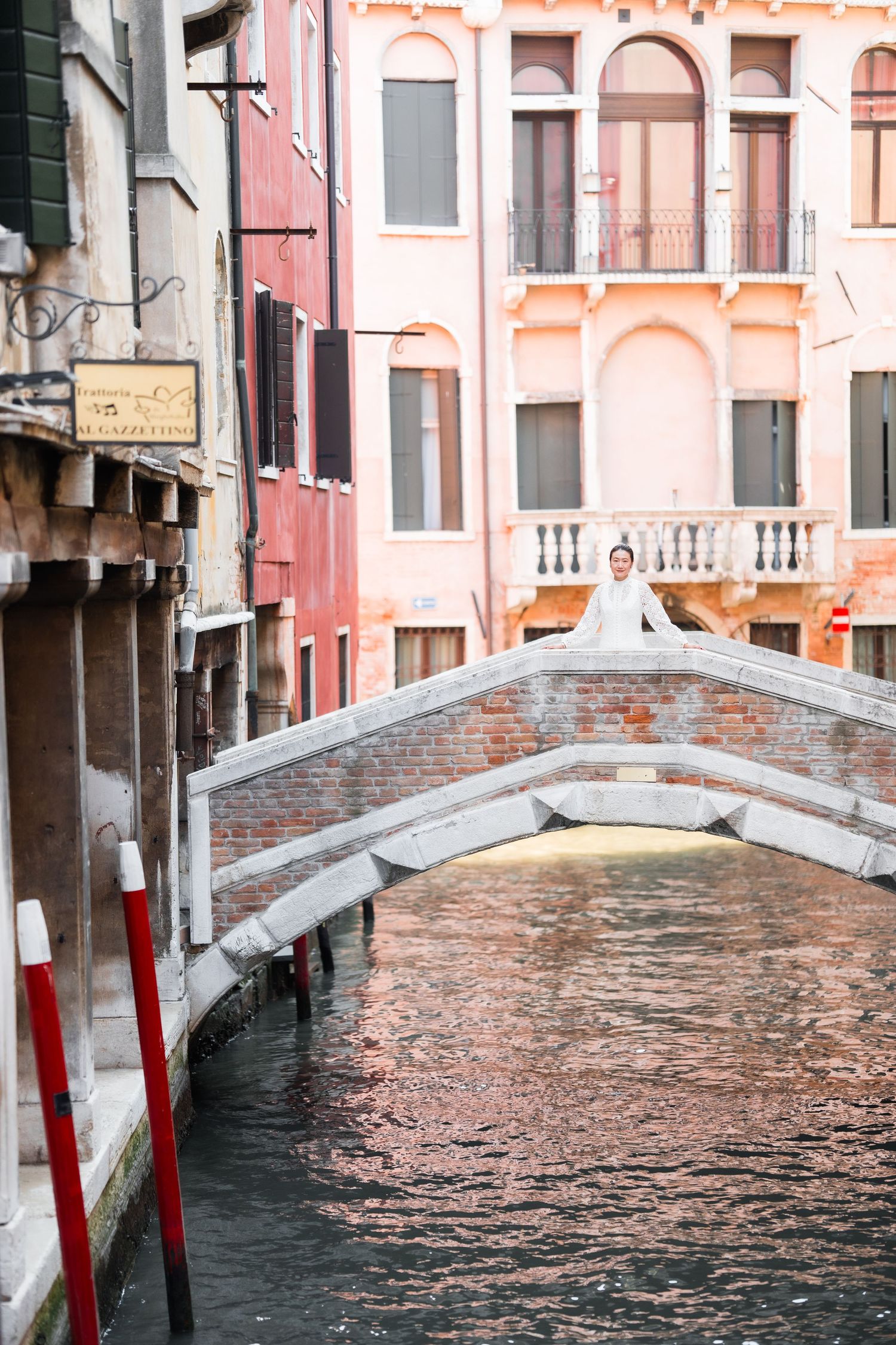 Bride in white wedding dress standing on a stone bridge over a Venice canal with colorful buildings.