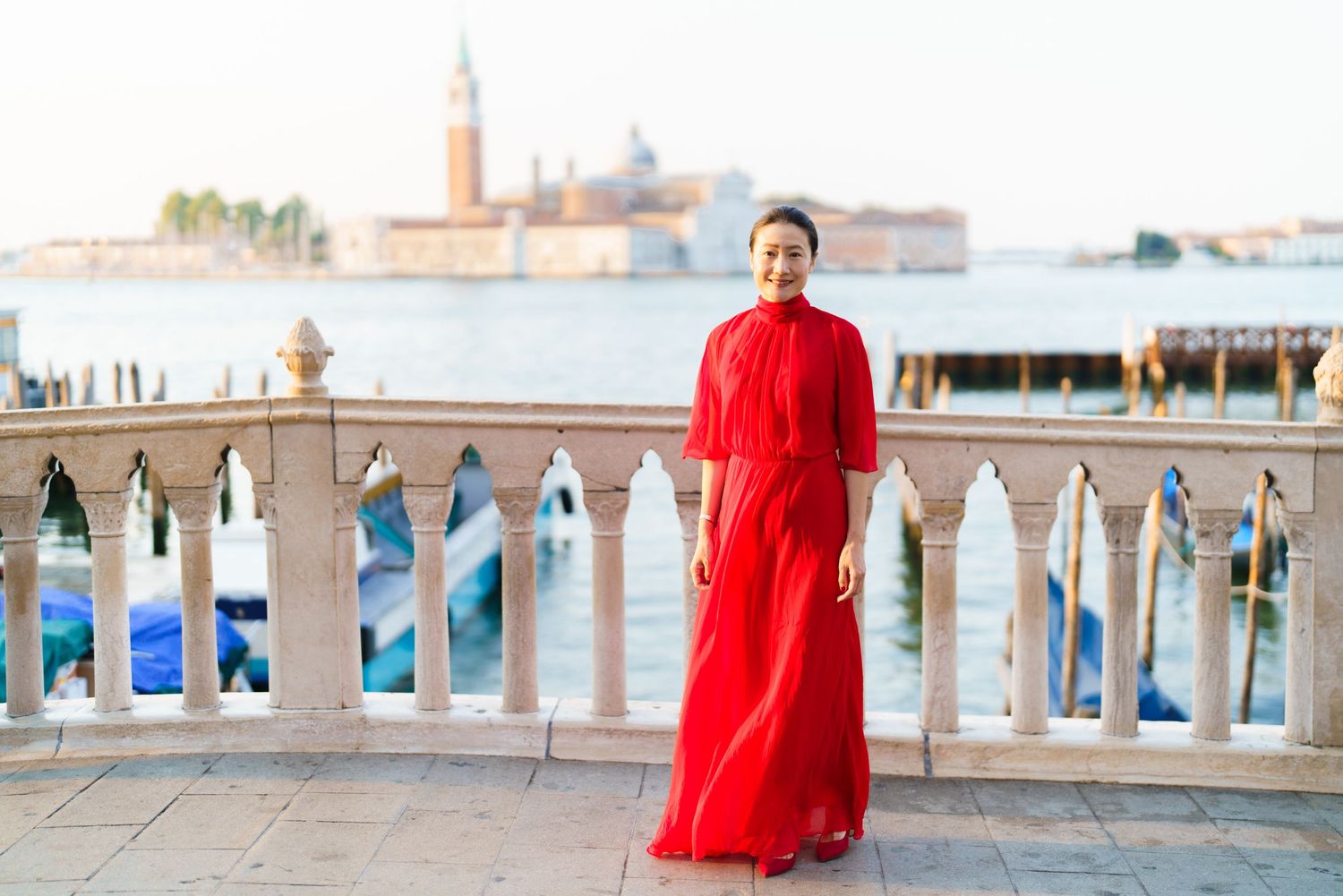 Woman in red dress standing on Venice waterfront terrace with gondolas and San Giorgio Maggiore island in background.