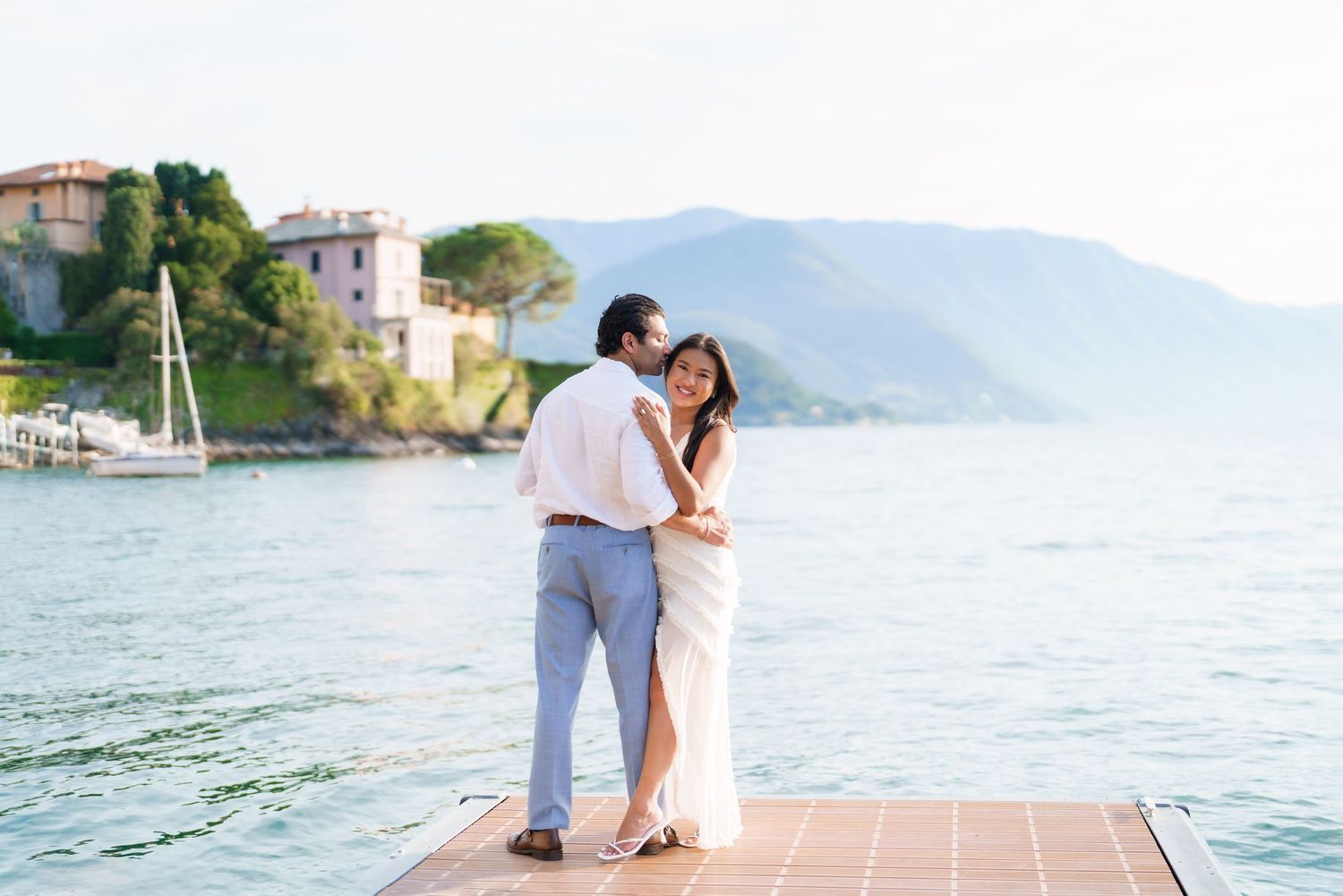Couple embraces on a dock with scenic lake and mountain views at golden hour.