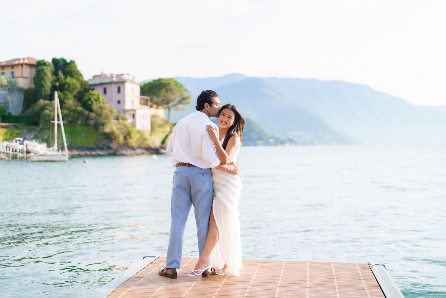 Couple embracing on a dock at Lake Como, Italy, with mountains and a historic villa in the background.