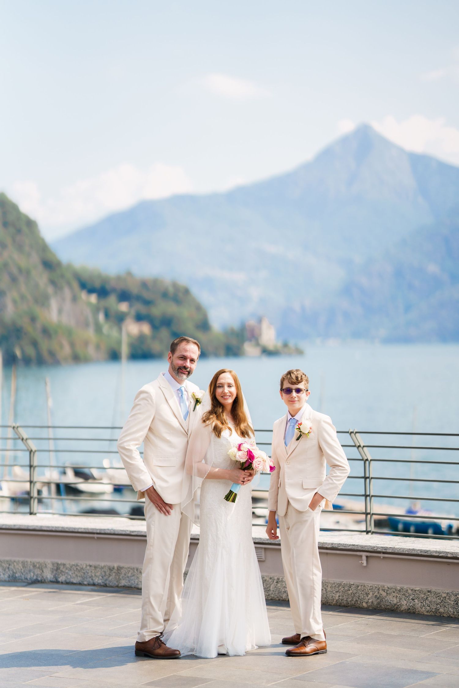 Bride and groom with guest posing in white suits on lakeside terrace with mountain backdrop.