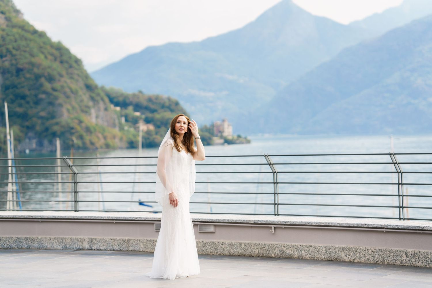 Bride in white gown poses on lakeside terrace with mountain backdrop at Lake Como, Italy.