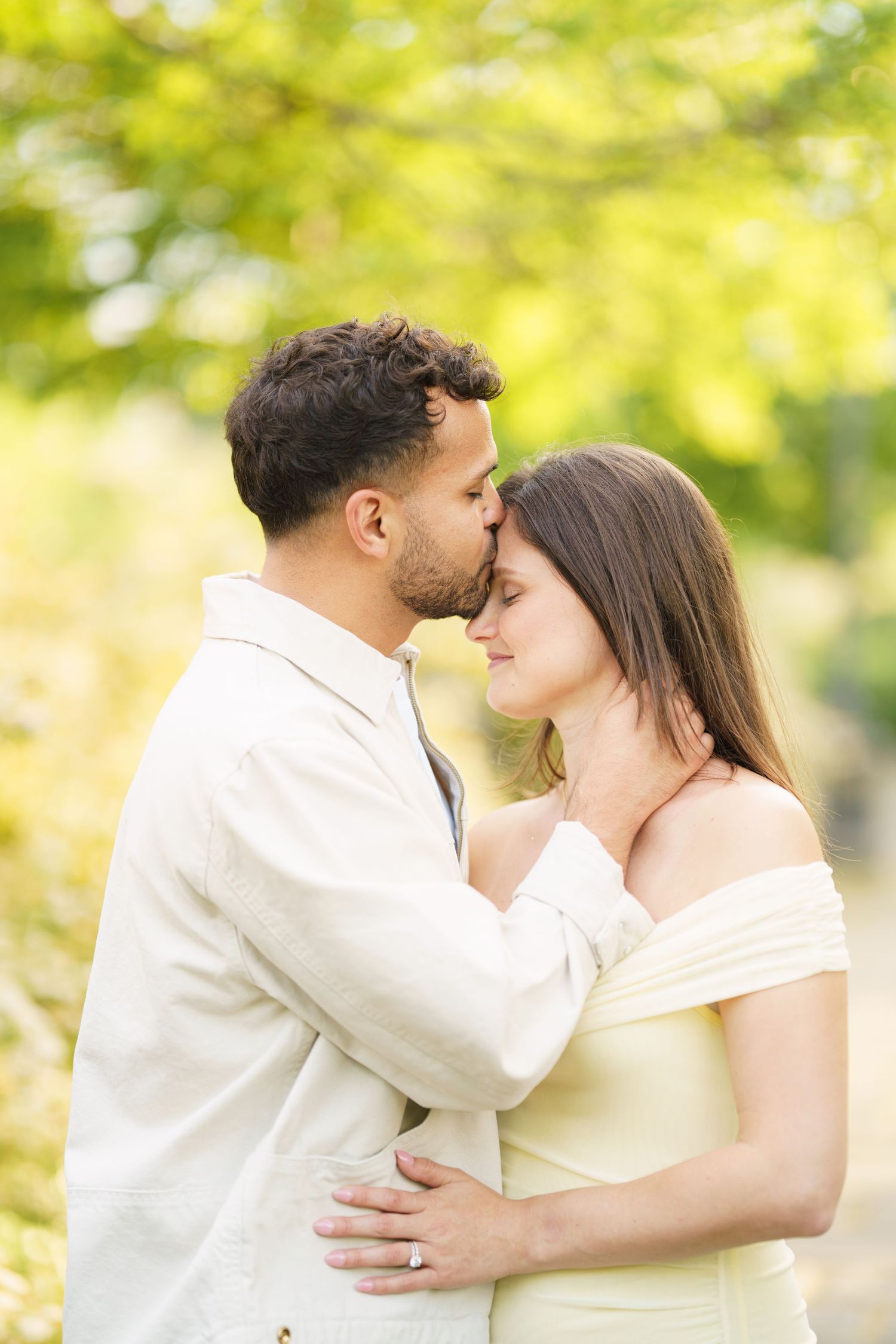 A couple shares a tender kiss outdoors, surrounded by lush green foliage, dressed in soft neutral tones.