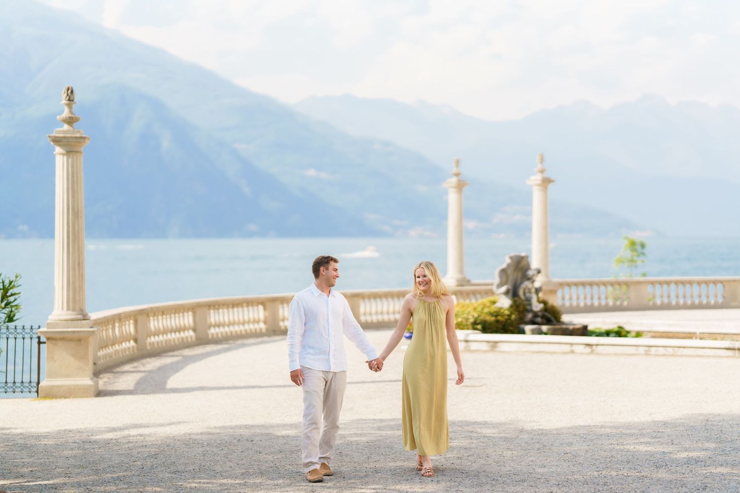 Couple holding hands on elegant lakeside terrace with mountain backdrop at Lake Como, Italy.