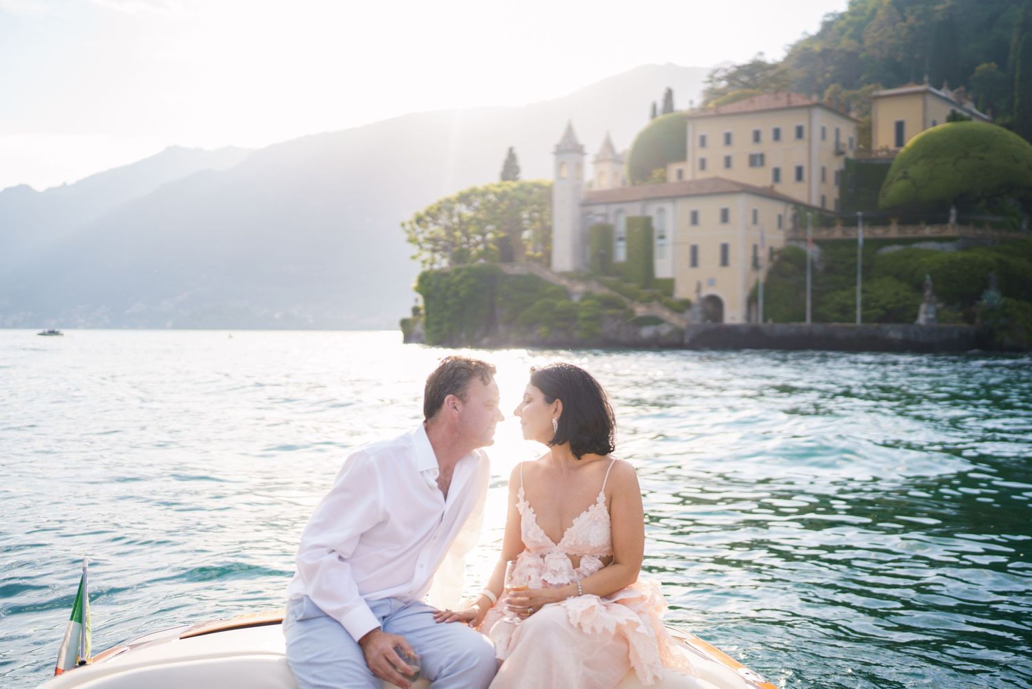 Couple sharing a romantic moment on a boat at Lake Como, Italy, with a sunlit villa and mountains in the background.