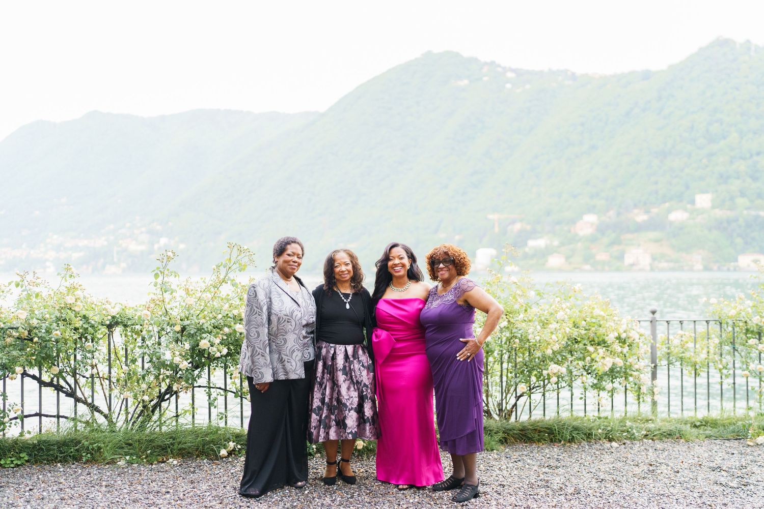Four women posing together outdoors with scenic mountain lake views and lush greenery in the background.
