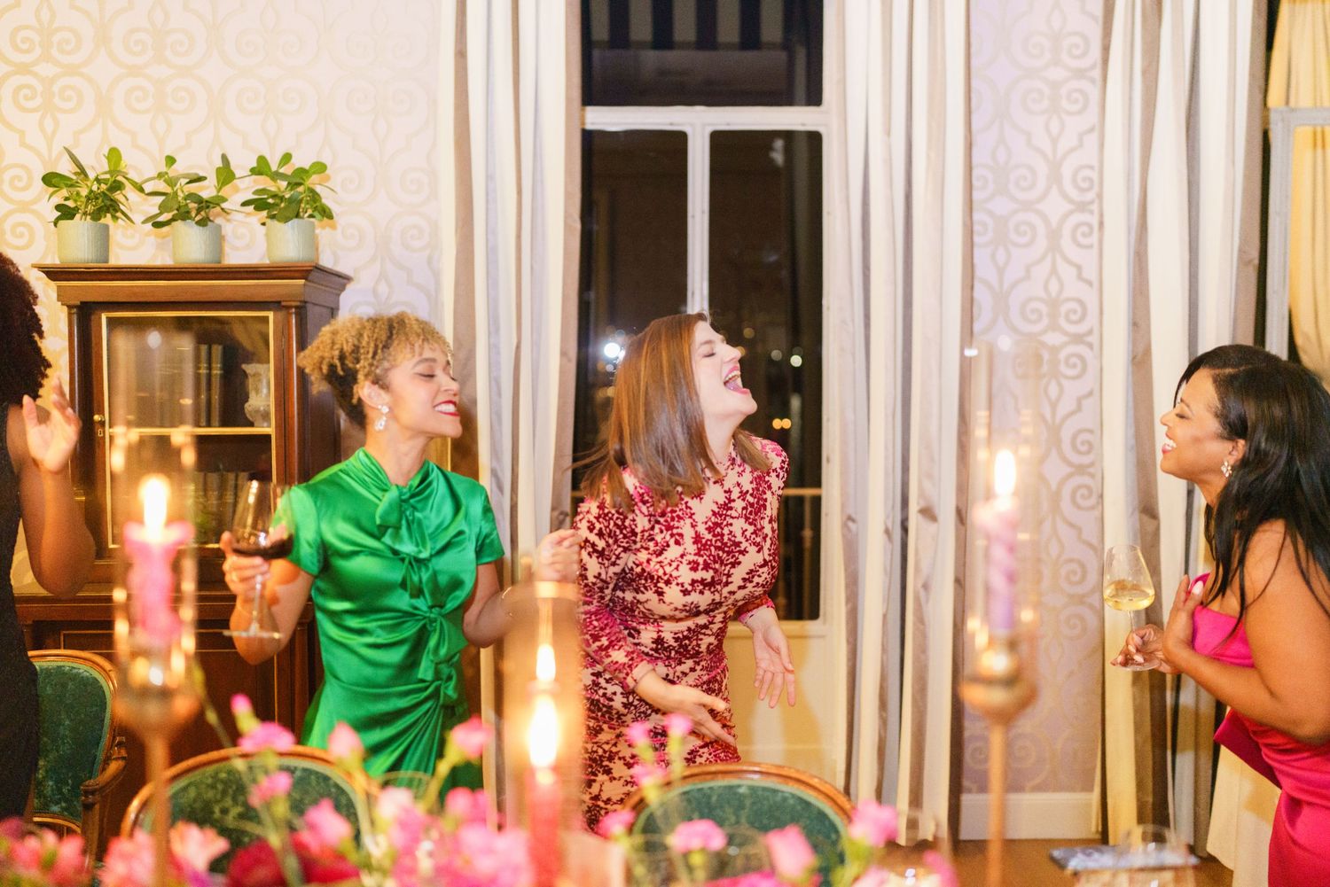 Women laughing and celebrating at an elegant candlelit dinner party with floral arrangements and champagne.