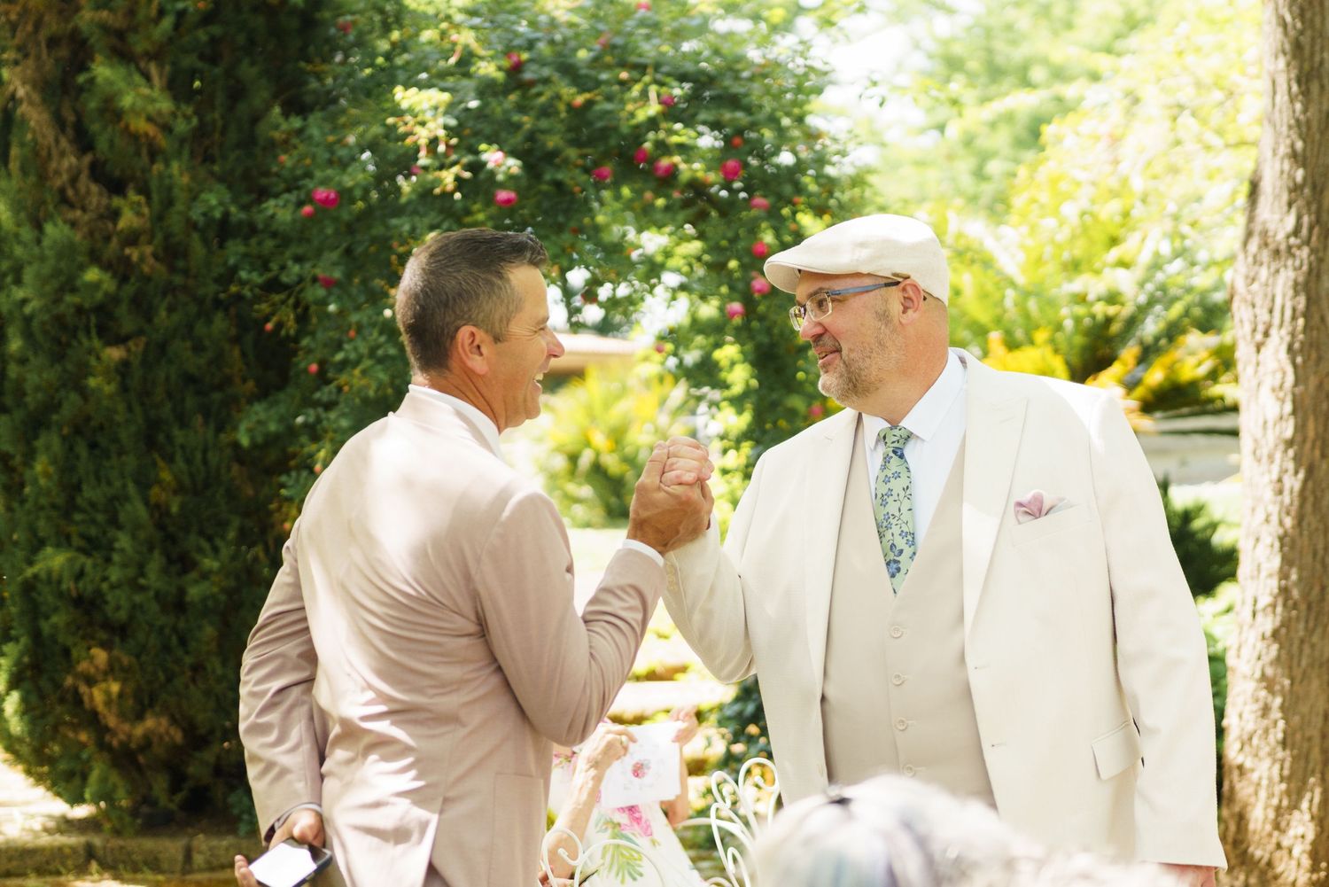 Two men in suits shaking hands and smiling at an outdoor wedding reception surrounded by lush green trees.