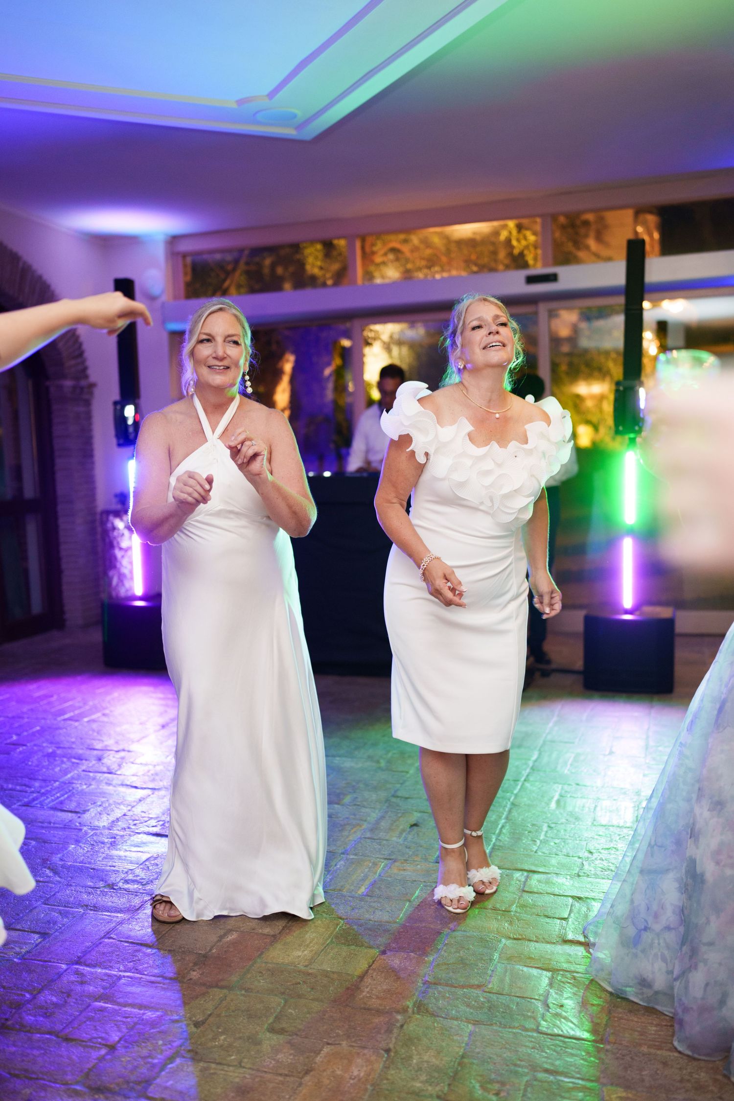 Two brides in white dresses dancing joyfully at their wedding reception under colorful purple and green lights.
