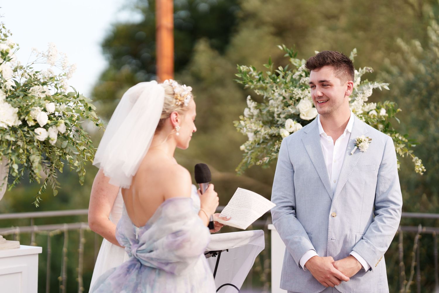 Bride reads vows to smiling groom in light gray suit at outdoor wedding ceremony with floral arch.
