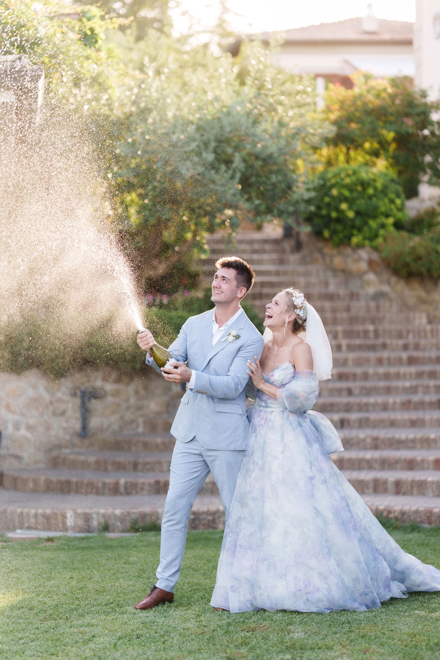 Joyful bride and groom popping champagne bottle outdoors on green lawn, wearing blue suit and floral wedding gown.