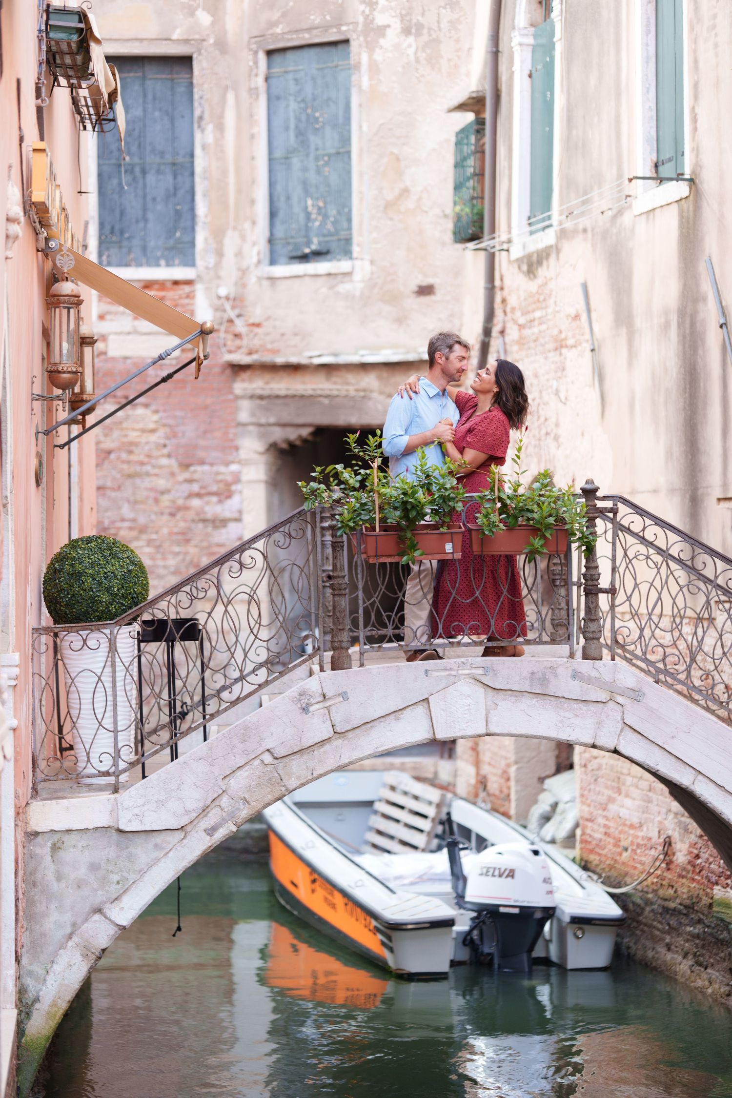Romantic couple embracing on a charming Venice canal bridge with boats and historic buildings in the background.