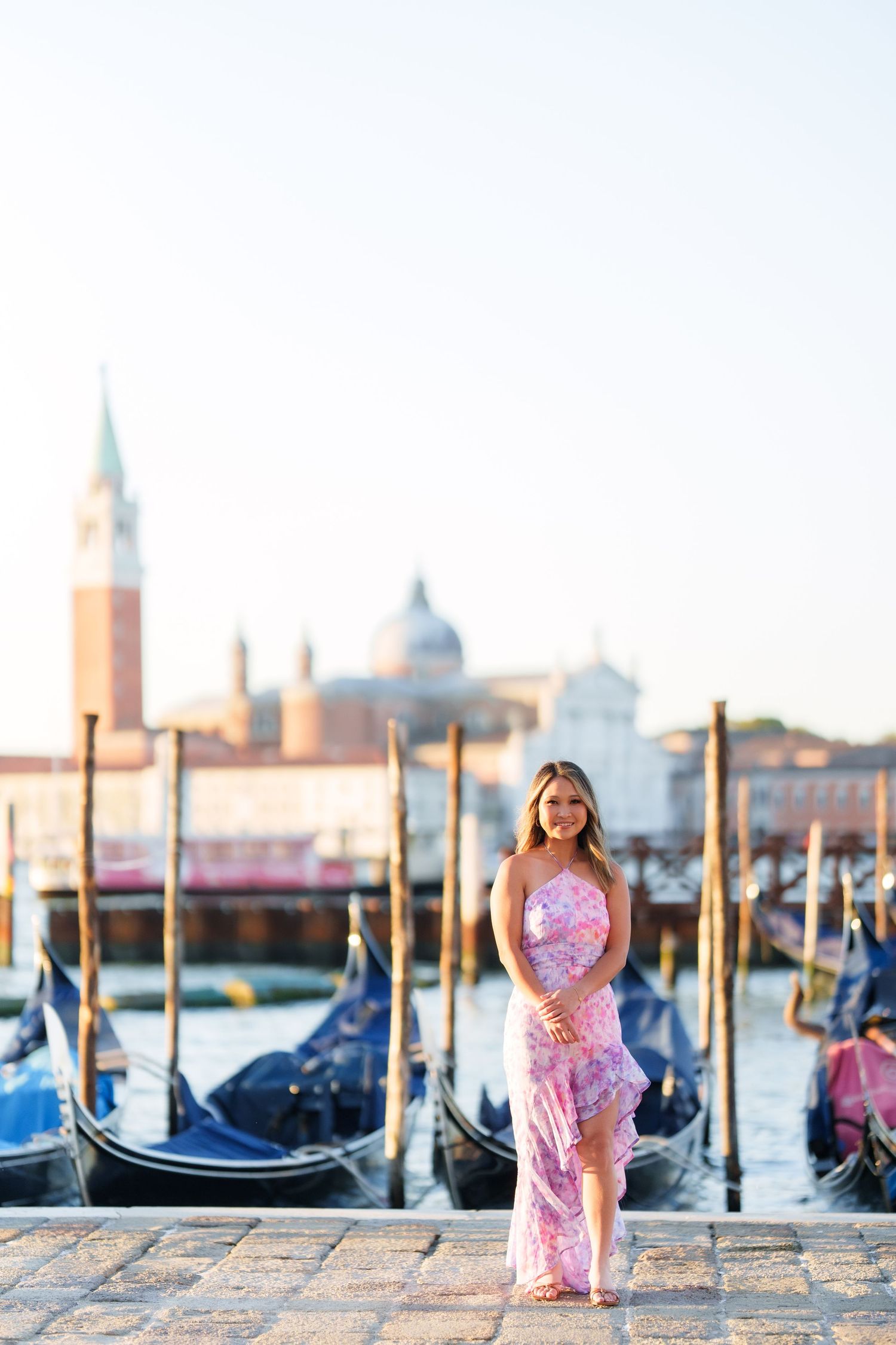 Woman in pink floral dress posing by Venice gondolas with San Giorgio Maggiore church in the background.