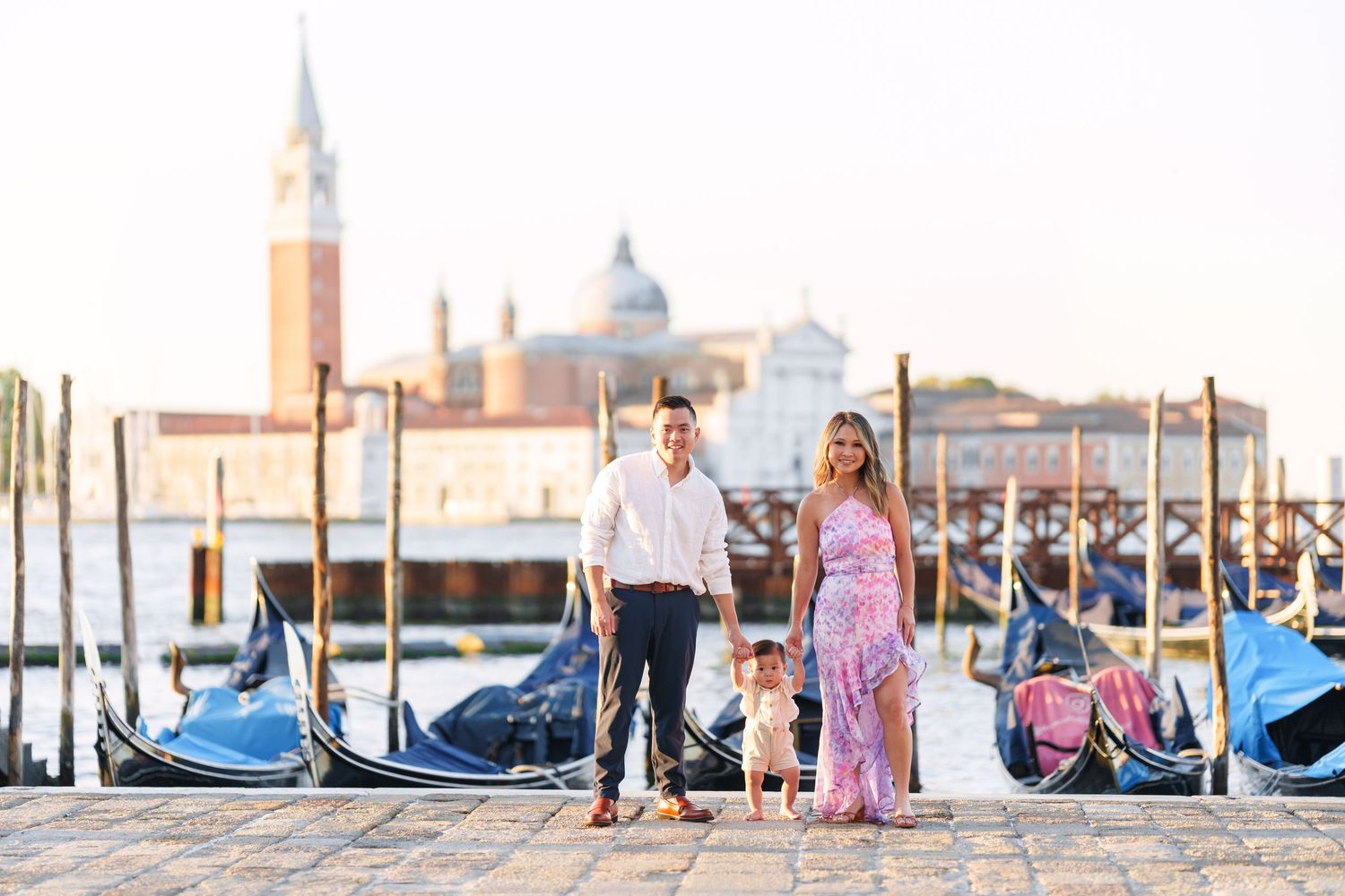 Family of three posing by gondolas on Venice waterfront with San Giorgio Maggiore church in background.