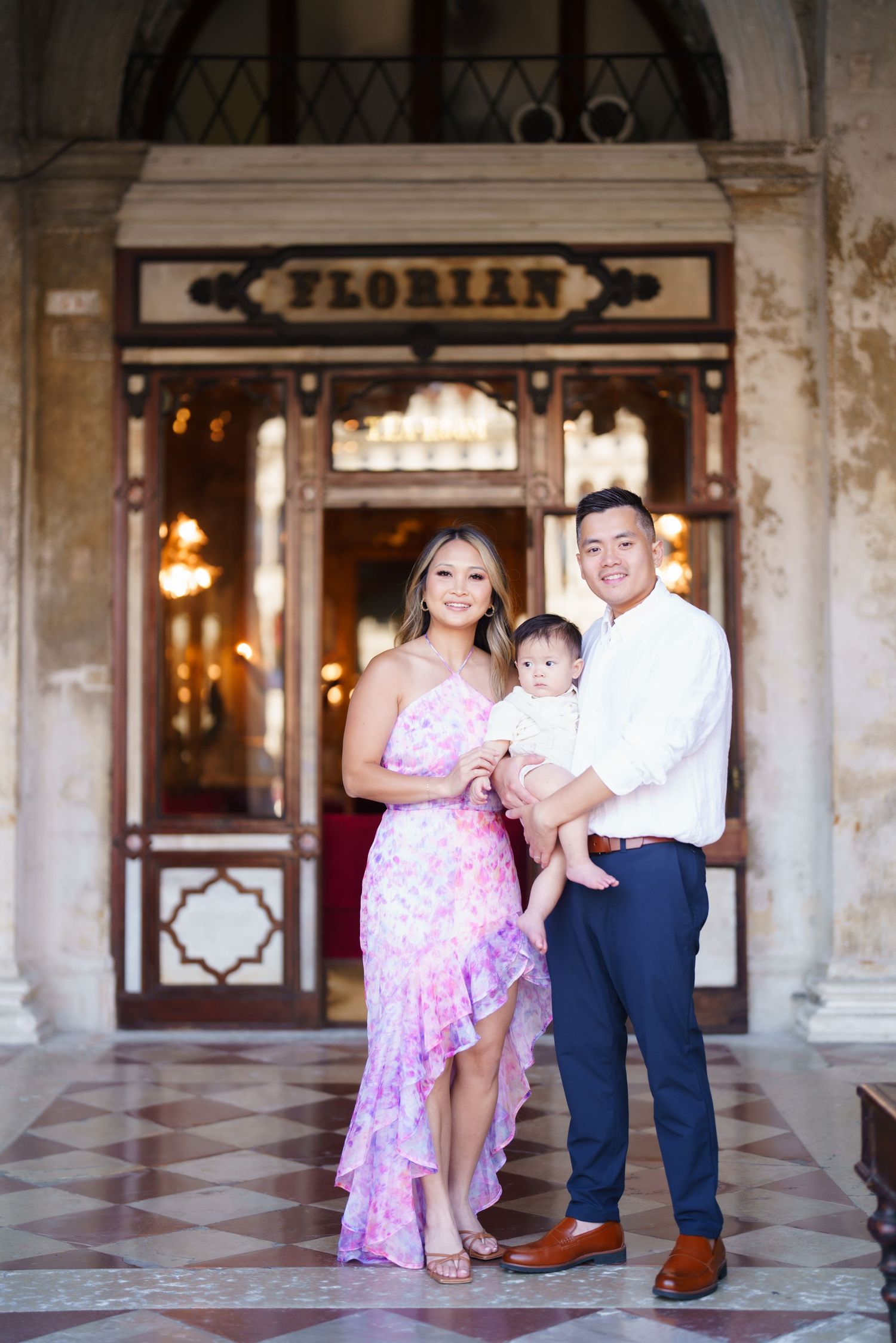 A smiling family of three poses in front of Caffe Florian in Venice, Italy, with a woman in a pink dress and a man holding their baby.