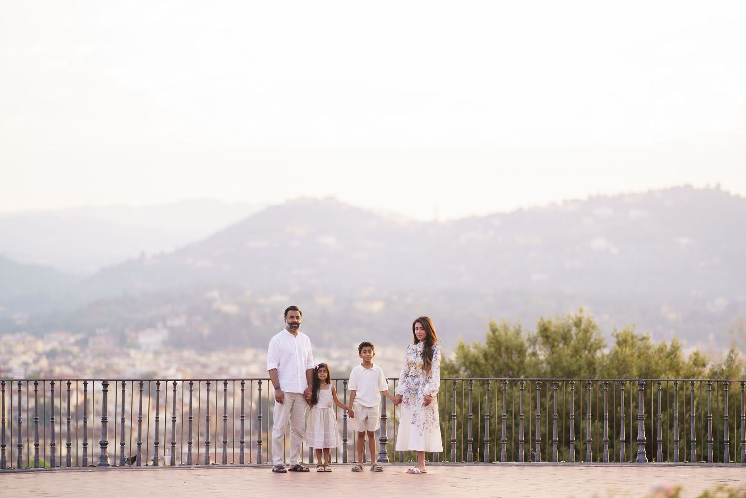 Family of four in white outfits posing on a scenic overlook with mountain views and lush greenery backdrop.