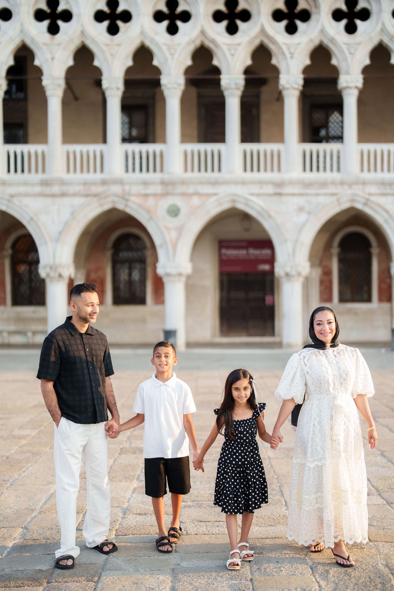 Family of four holding hands in front of Doge's Palace in Venice, Italy, dressed in elegant black and white outfits.