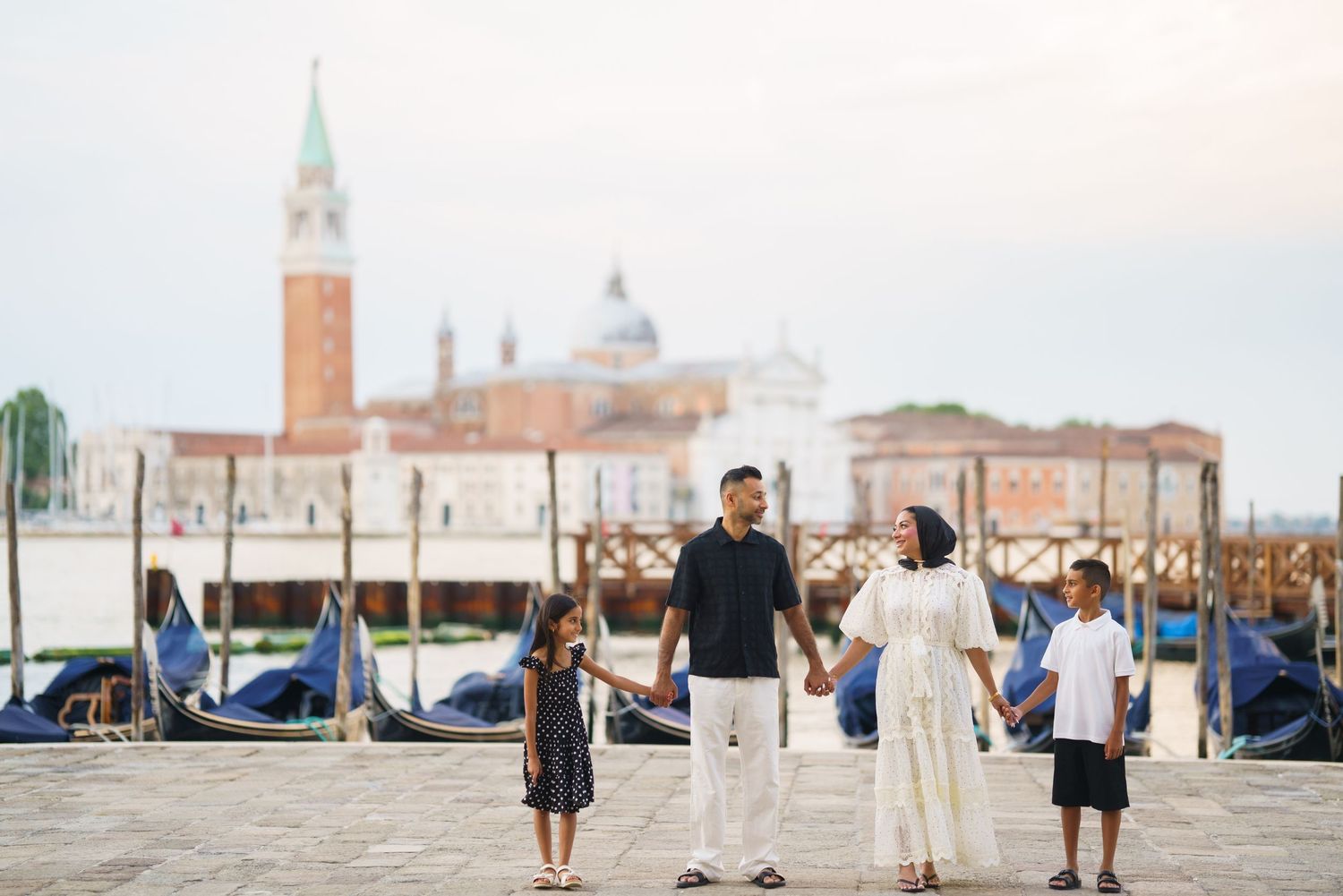 Family of four holding hands on Venice waterfront with gondolas and San Giorgio Maggiore church in background.