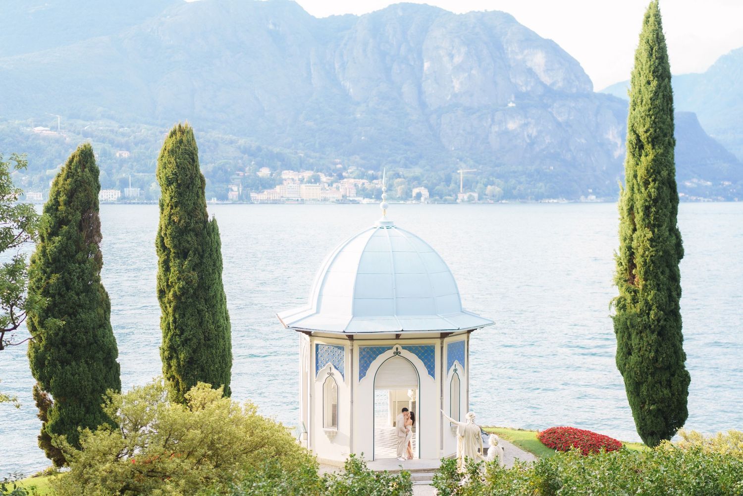 Scenic lakeside view with cypress trees and a white church building against mountainous backdrop in Italy.