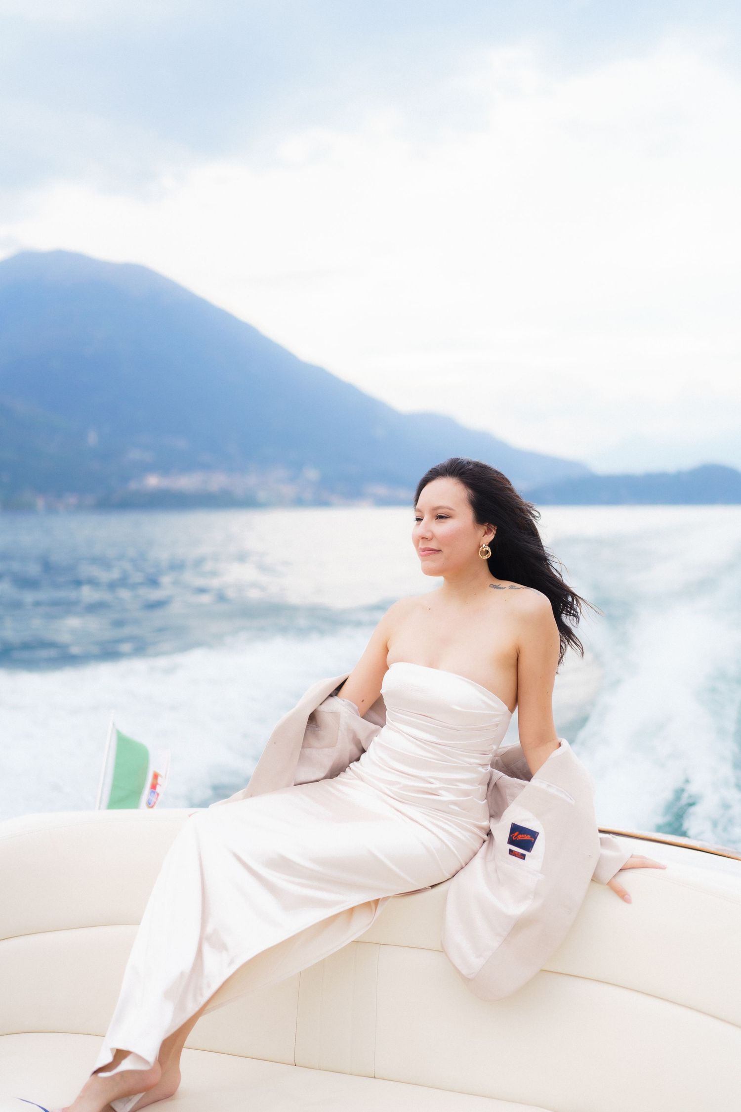 Figure in white dress relaxes on boat deck with mountains and lake in background on sunny day.
