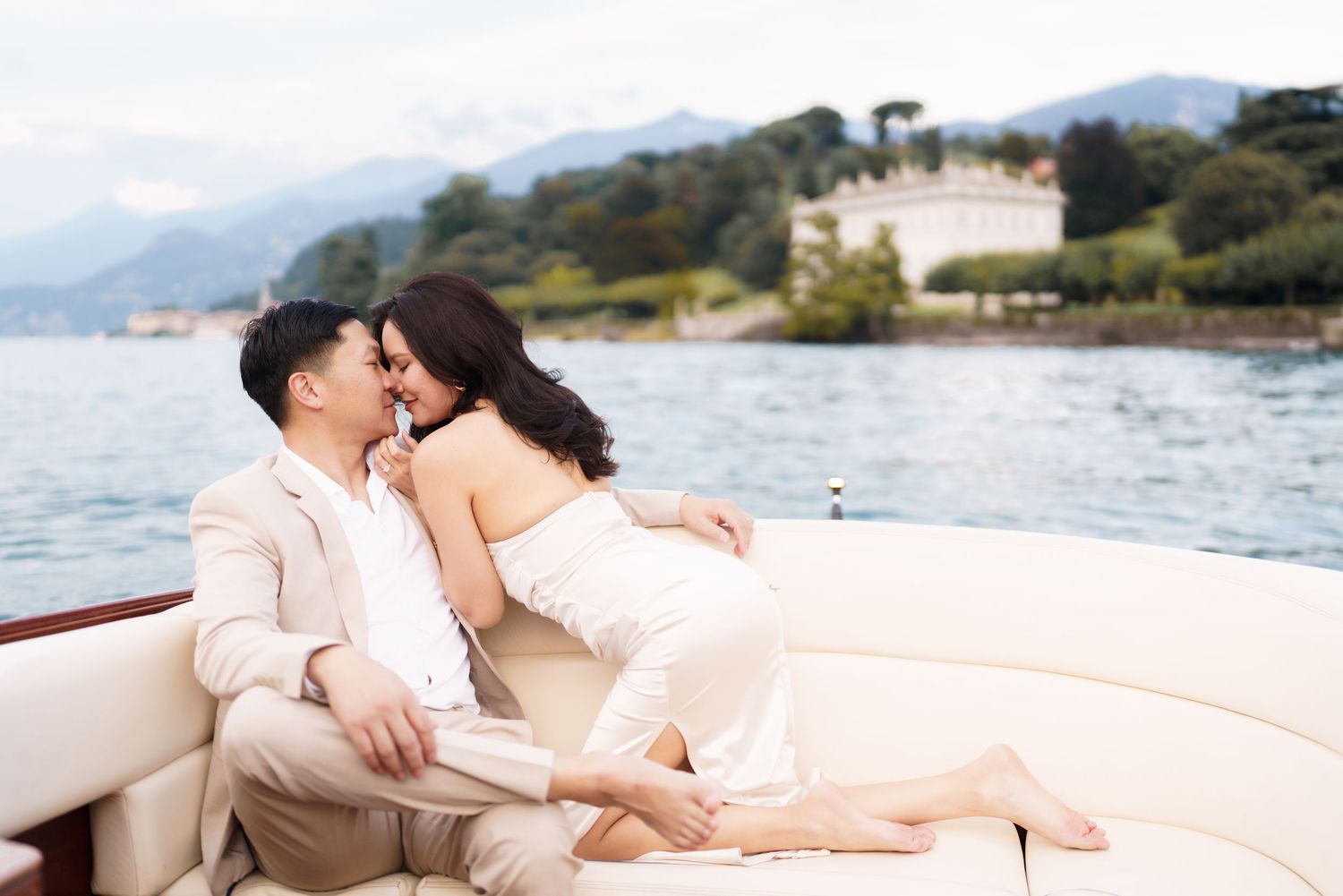 A couple shares a romantic moment while relaxing on a beige cushioned boat deck overlooking a scenic waterfront.