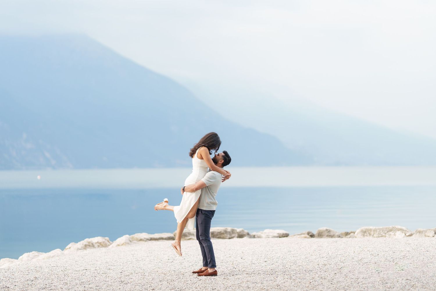 Couple embraces on a scenic mountain beach during a romantic photoshoot with misty peaks in the background.