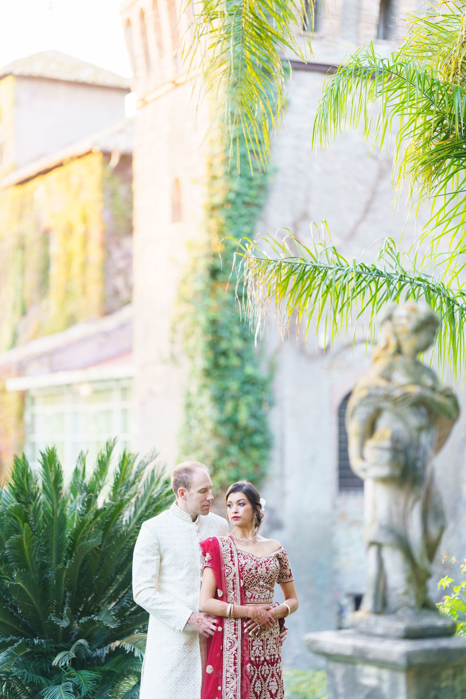 A couple embraces near stone statues and palm trees at a historic venue during golden hour.