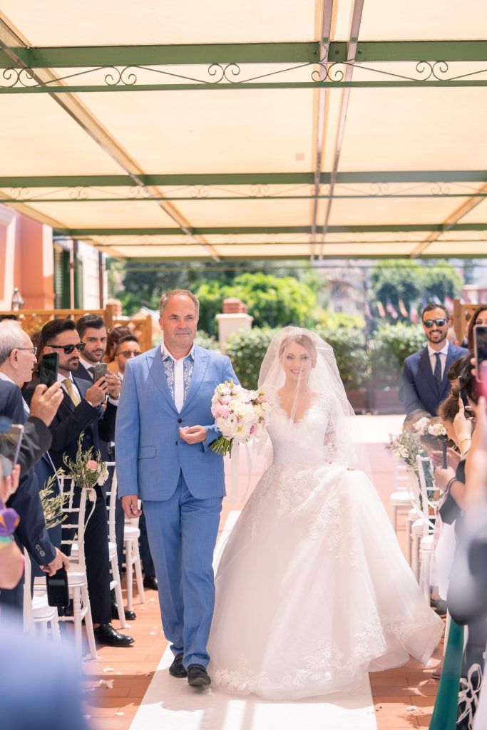 Wedding ceremony procession on covered terrace with guests capturing photos on phones.