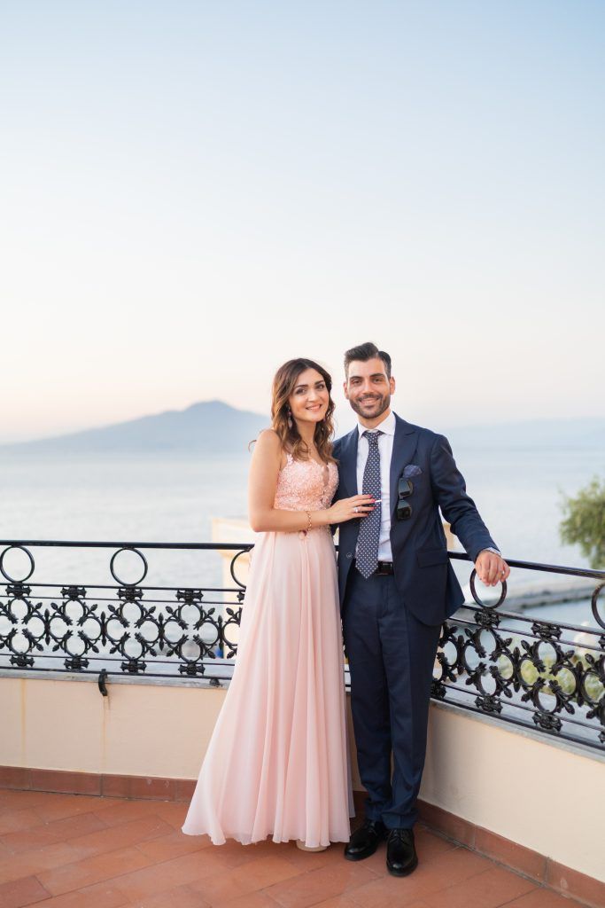 Elegant couple posing on a seaside terrace with ornate railings at sunset.