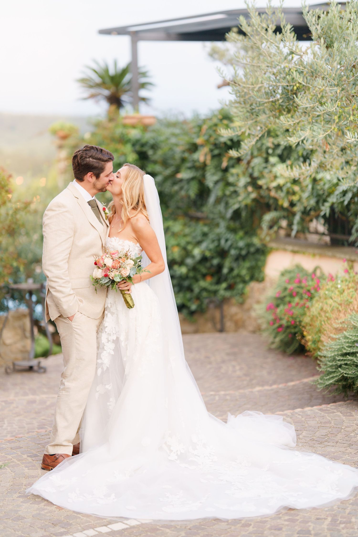 A romantic wedding couple embraces on a terrace with lush greenery and flowers in the background.