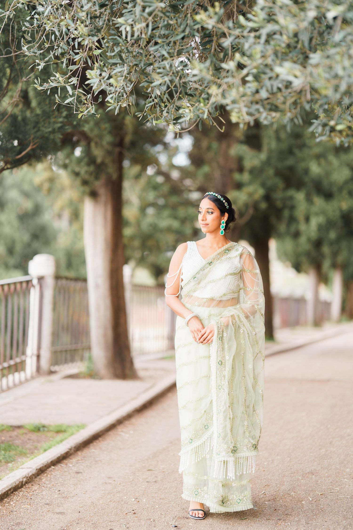 A person in a light green traditional dress poses gracefully along a tree-lined path in an outdoor setting.