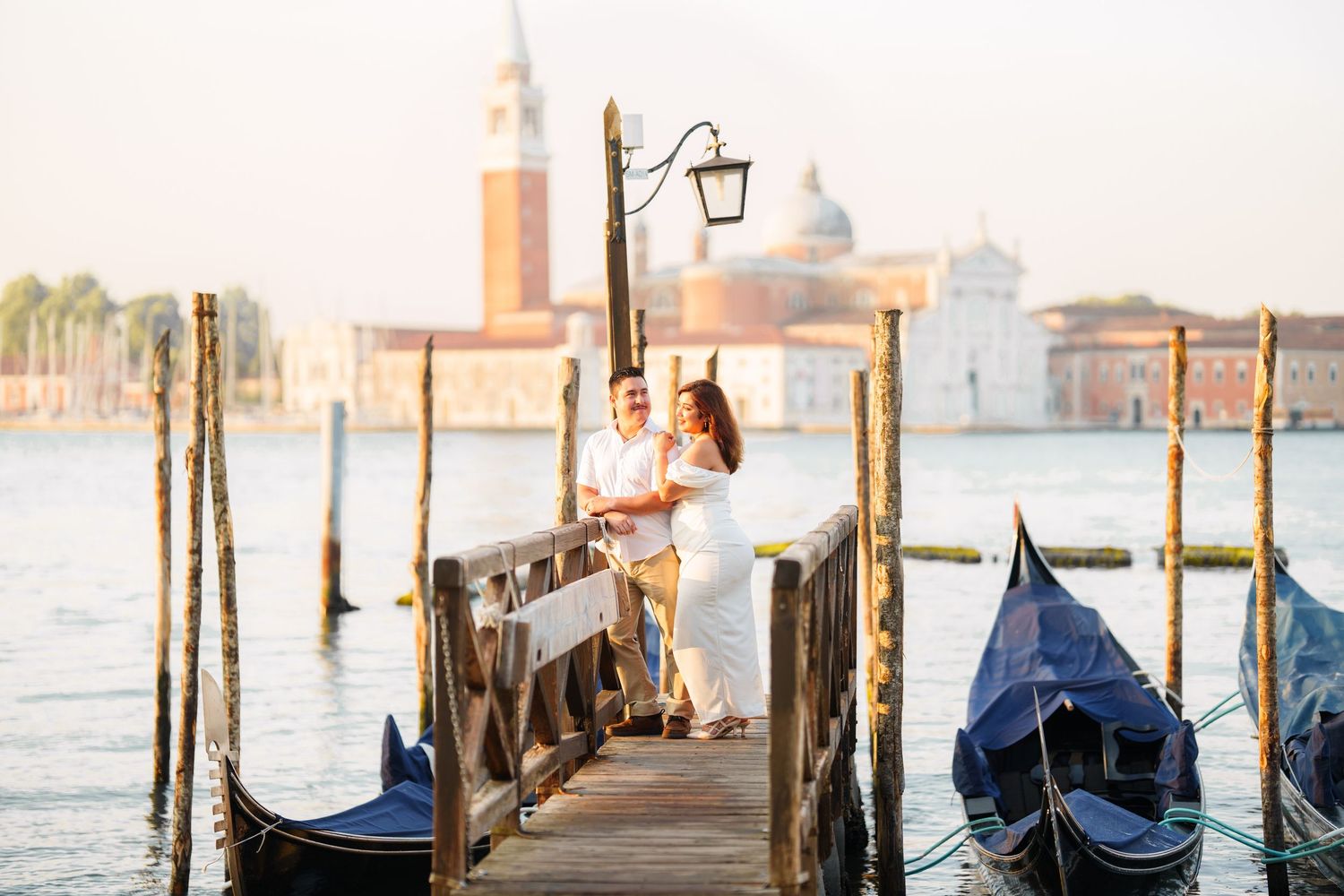 photographer venice A row of traditional Venetian gondolas moored along a misty canal with historic buildings and lamp posts at sunset.