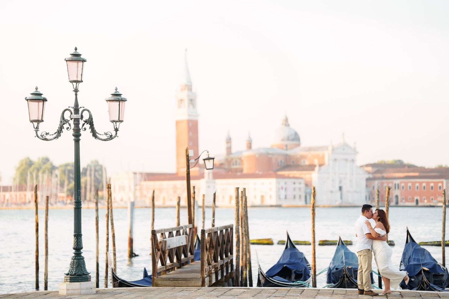 A romantic couple embraces near ornate lampposts along Venice's Grand Canal with covered gondolas and San Giorgio church visible.