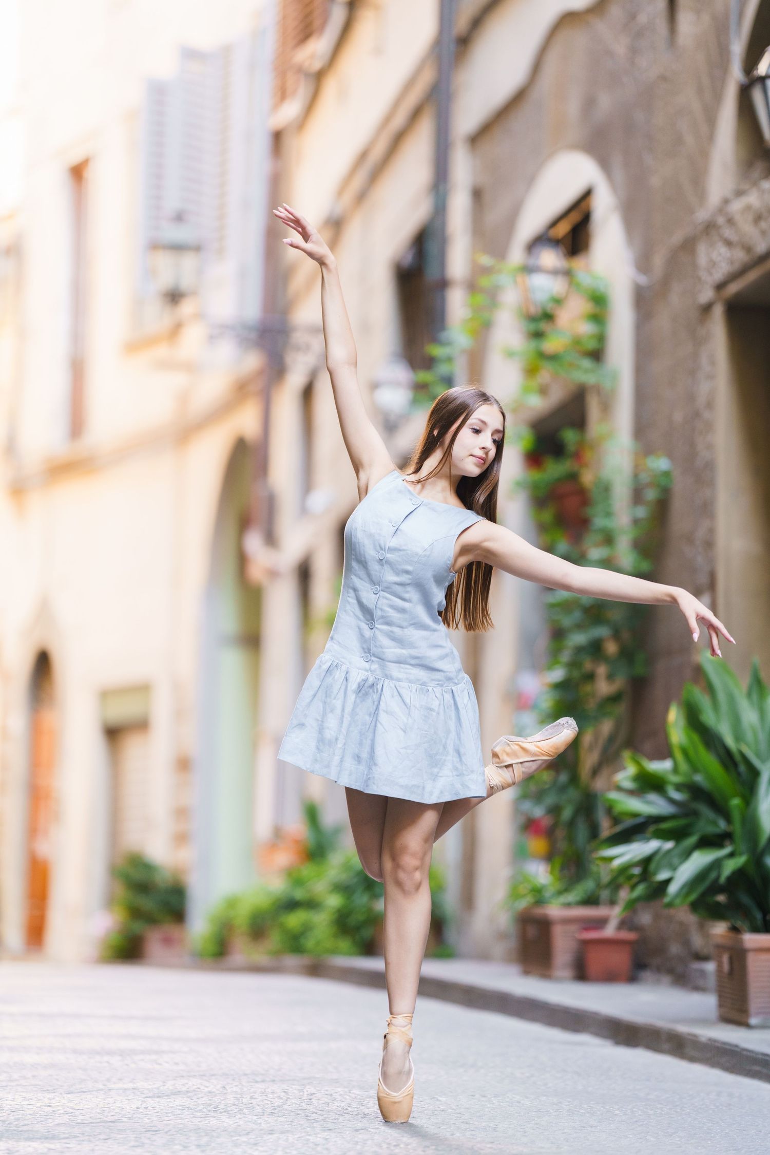 Graceful ballet dancer performs elegant poses in a charming European street setting with stone buildings and greenery.