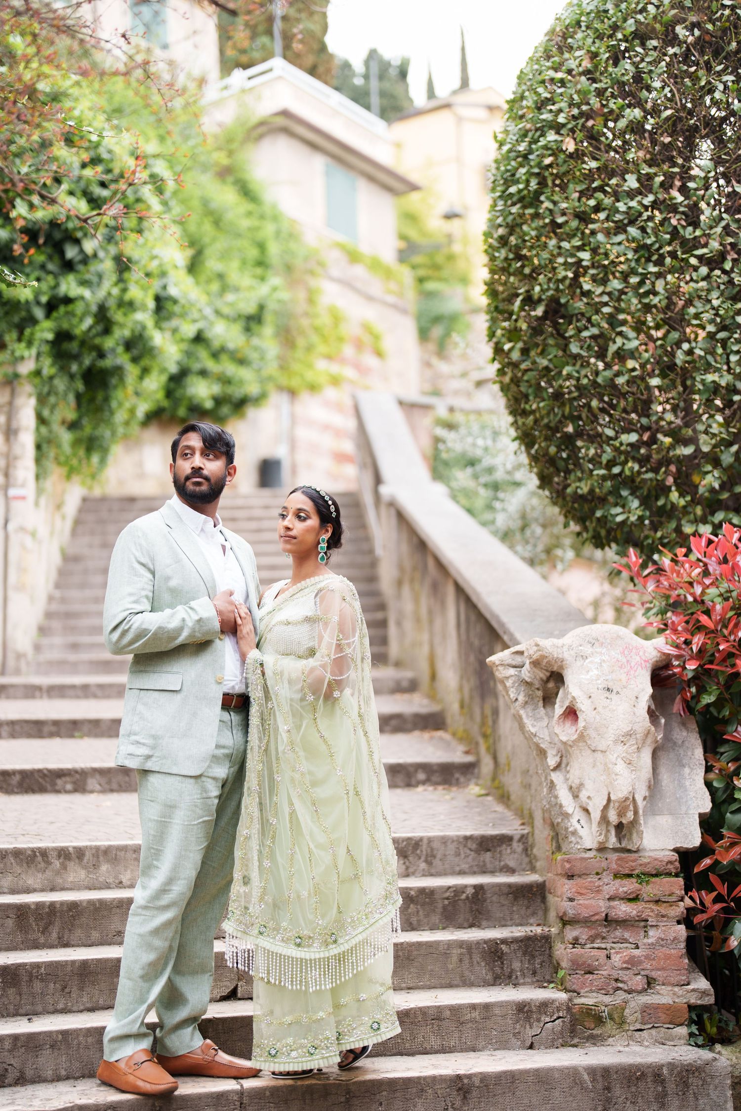 A couple in traditional South Asian wedding attire pose together on stone steps surrounded by greenery and flowers.
