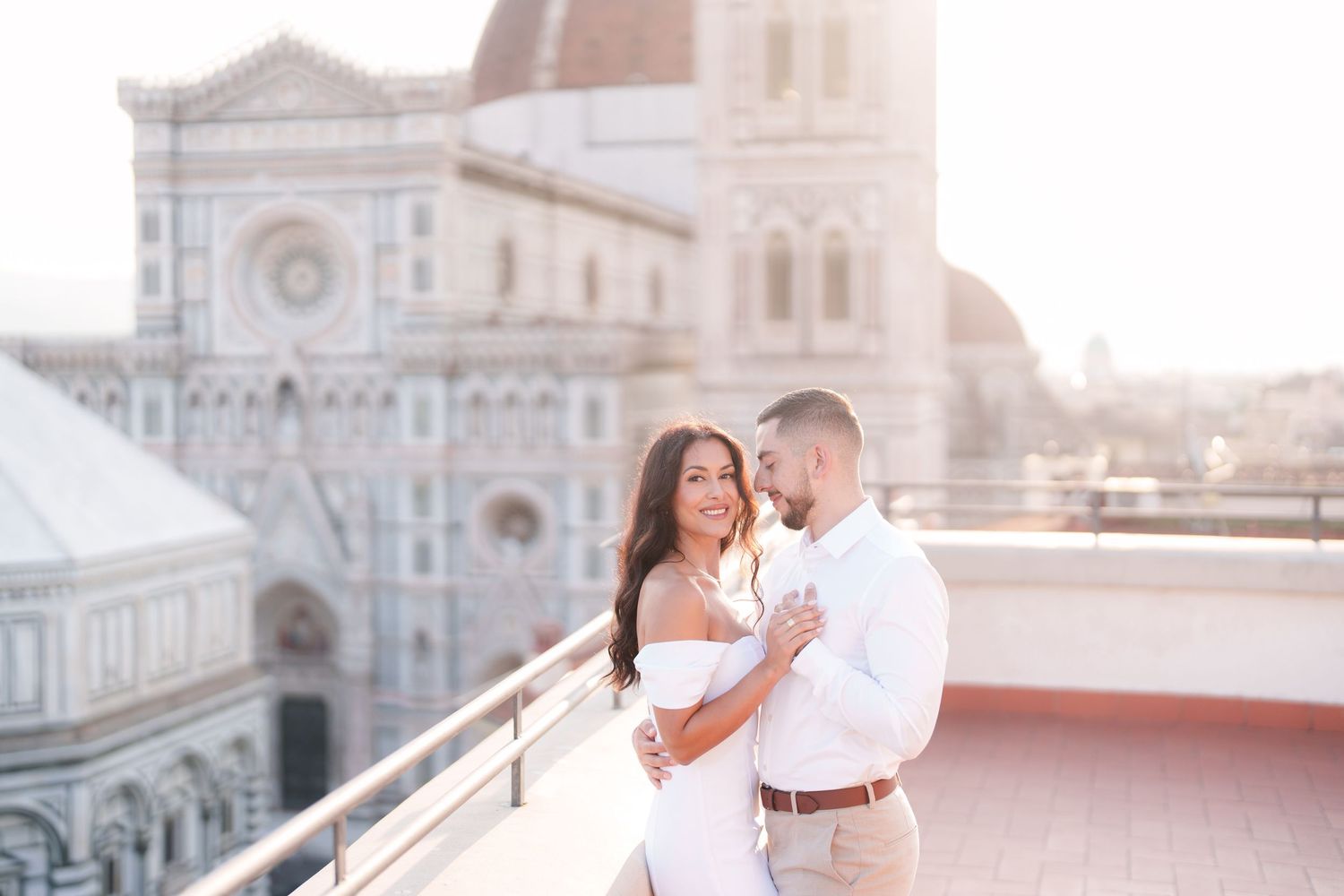 A couple in white outfits embraces romantically on a rooftop terrace overlooking Florence Cathedral during sunset.