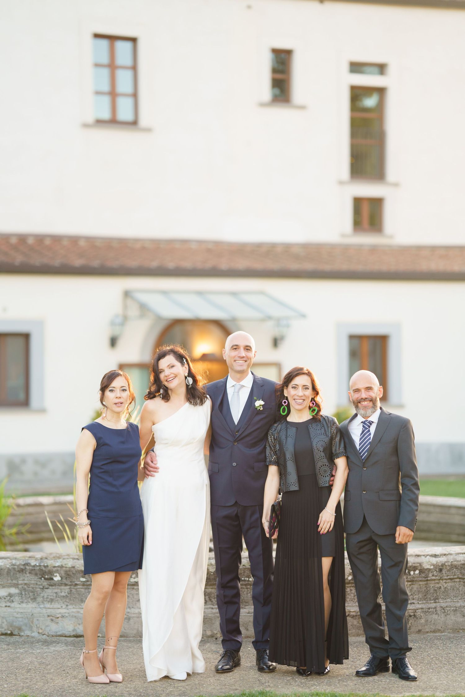 Small wedding party group in formal navy and white attire posed outside building.
