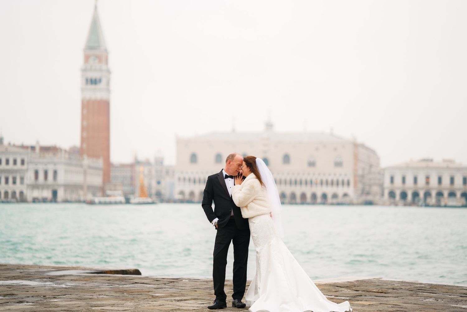 Couple in formal attire embrace while standing on a dock overlooking Venice's iconic waterfront and architecture.