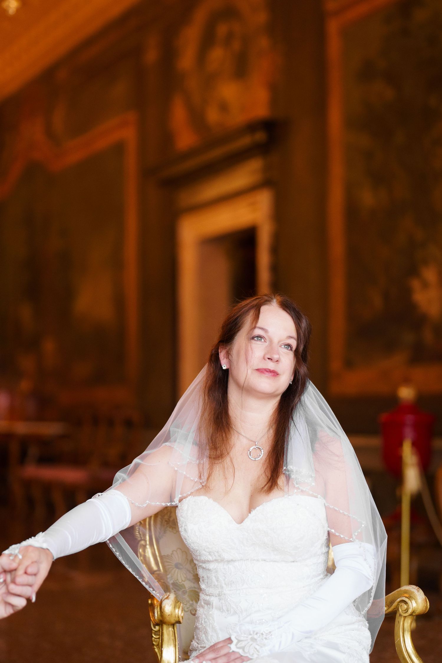 A bride in a white lace wedding dress and veil poses in a warm-lit room with ornate gold decor.