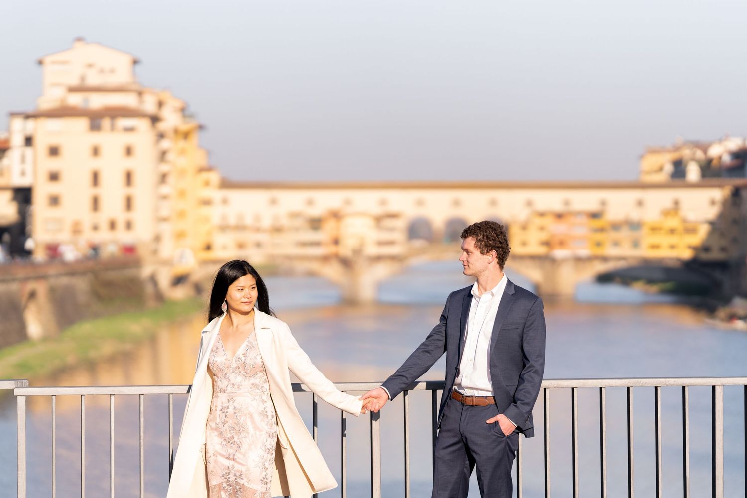 Couple shares romantic moment on bridge railing with Florence's Ponte Vecchio and Arno River in golden sunset light.