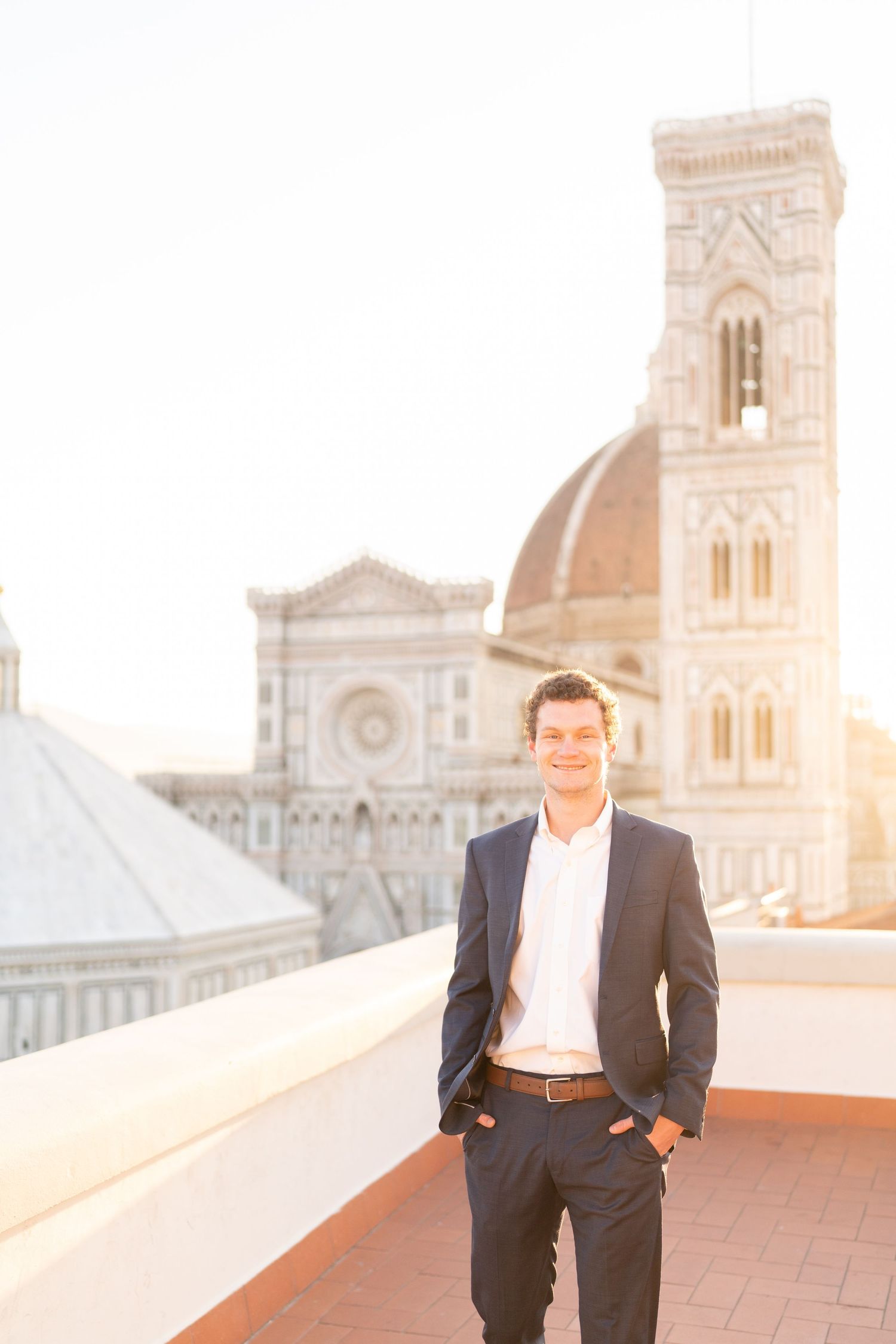 A man in a dark suit poses on a rooftop terrace with ornate architecture and golden sunlight in the background.