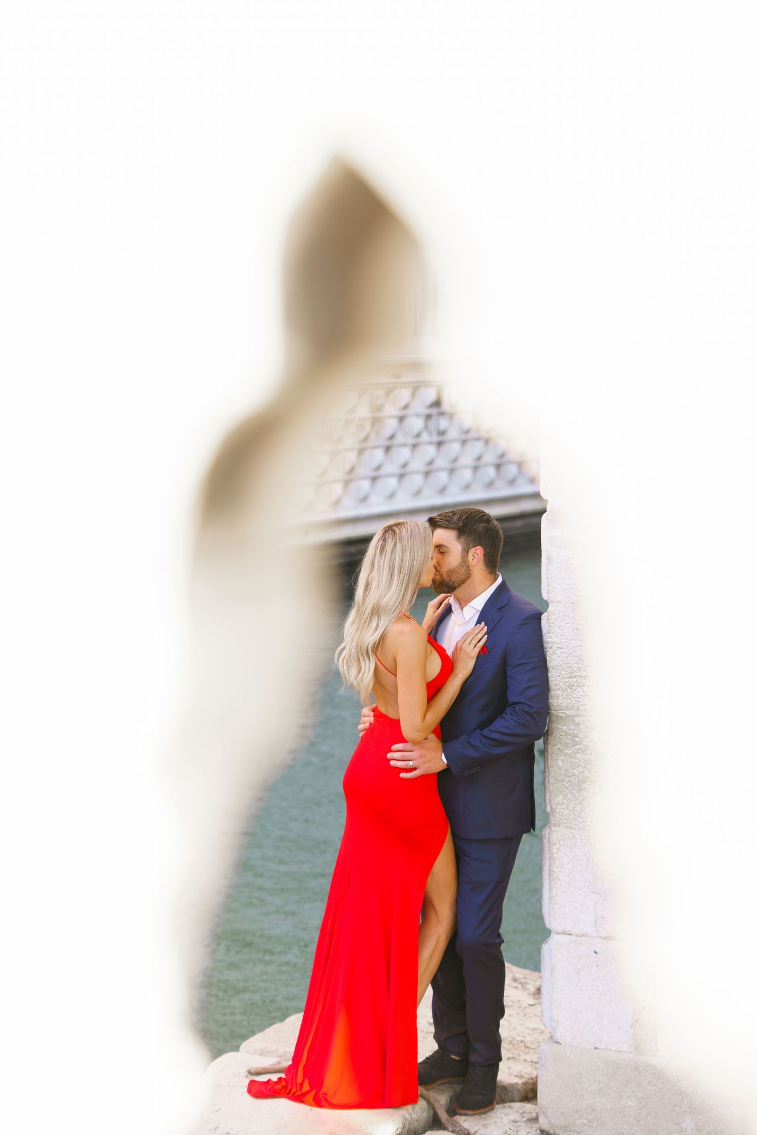 A romantic couple embraces by the water, with the person in a vibrant red dress and their partner in a navy suit.