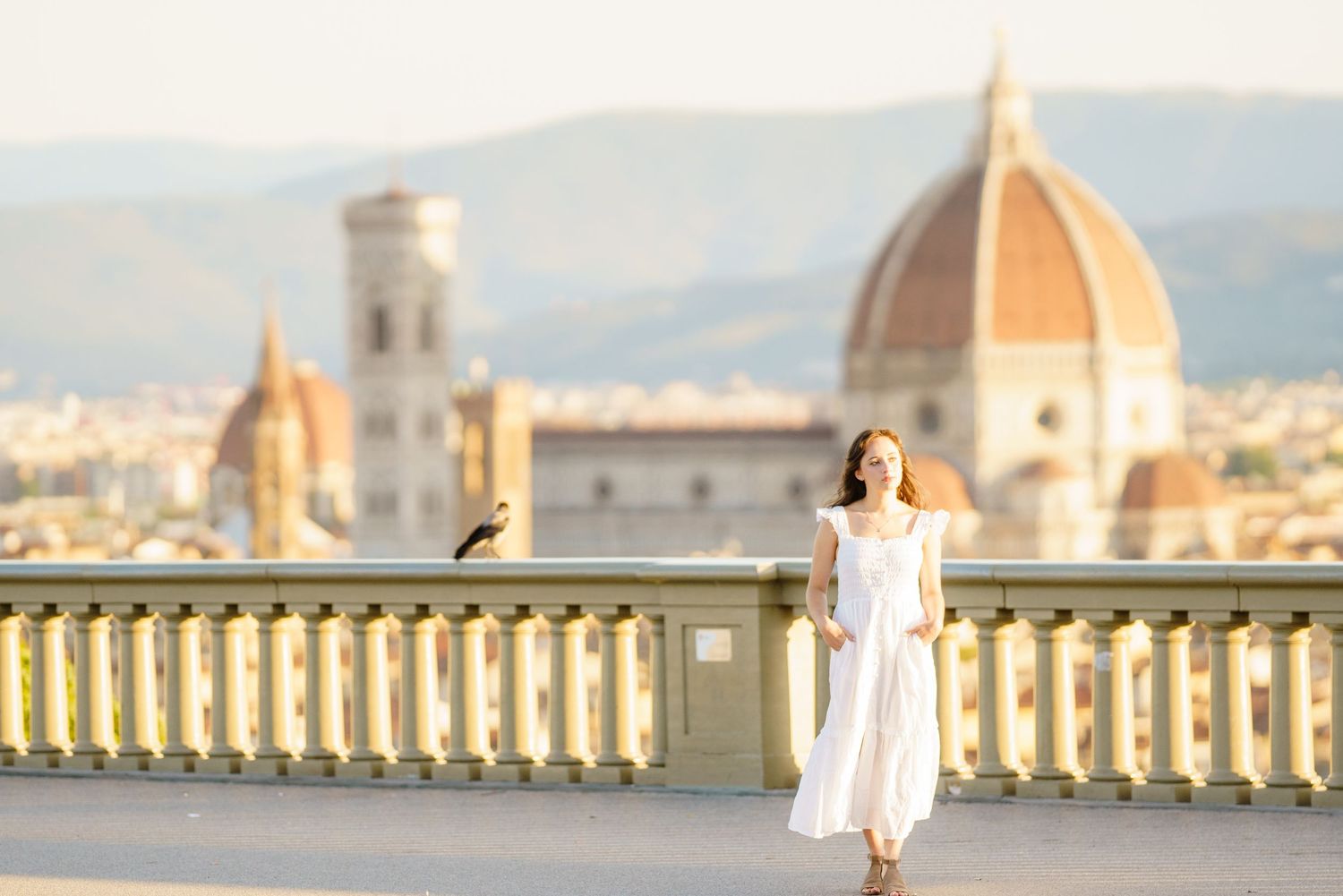 A person in white dress stands on a balcony overlooking the historic skyline of Florence, Italy at sunset.