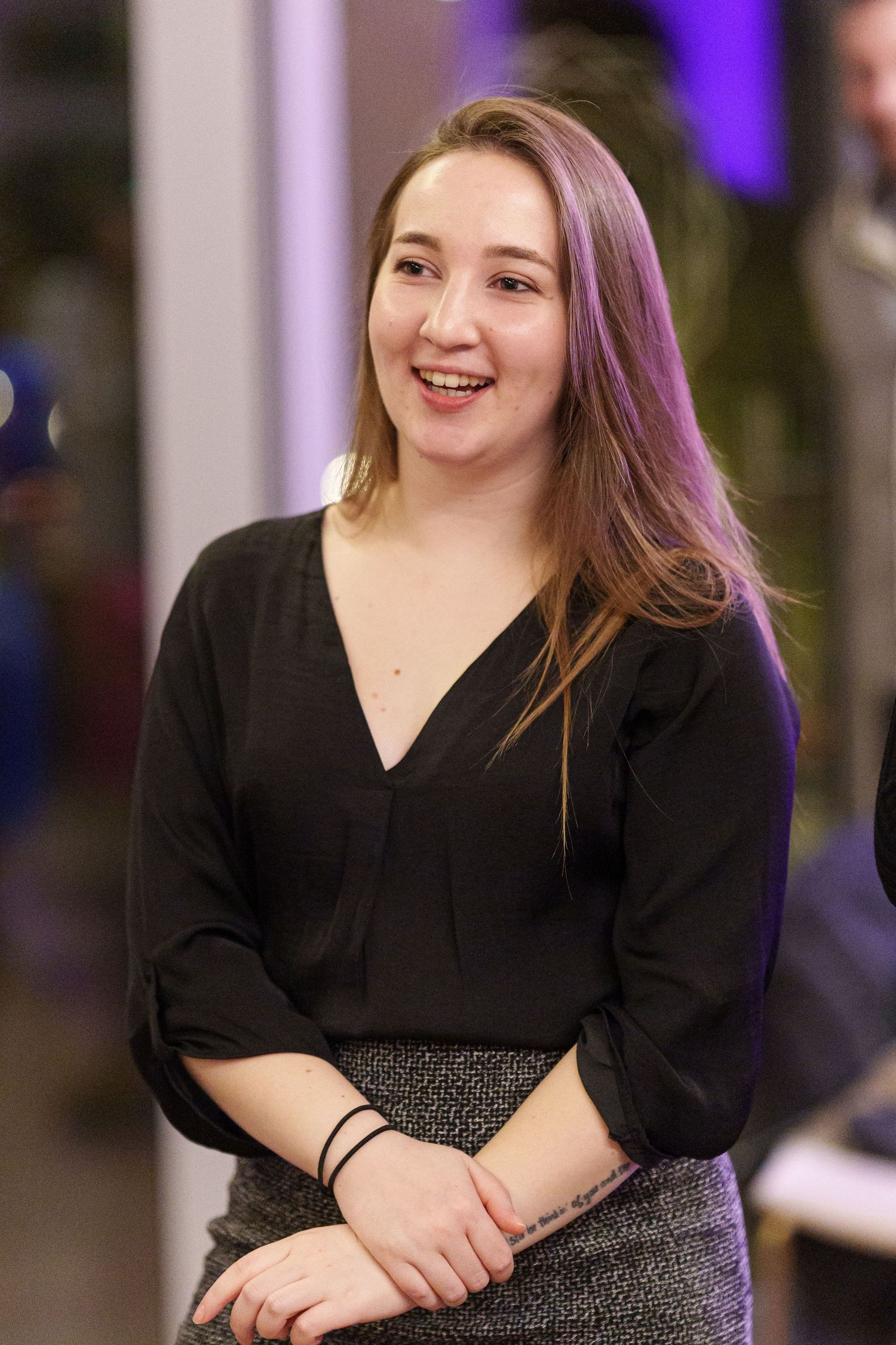 Smiling young woman with long brown hair wearing a black blouse and patterned skirt at an indoor event.